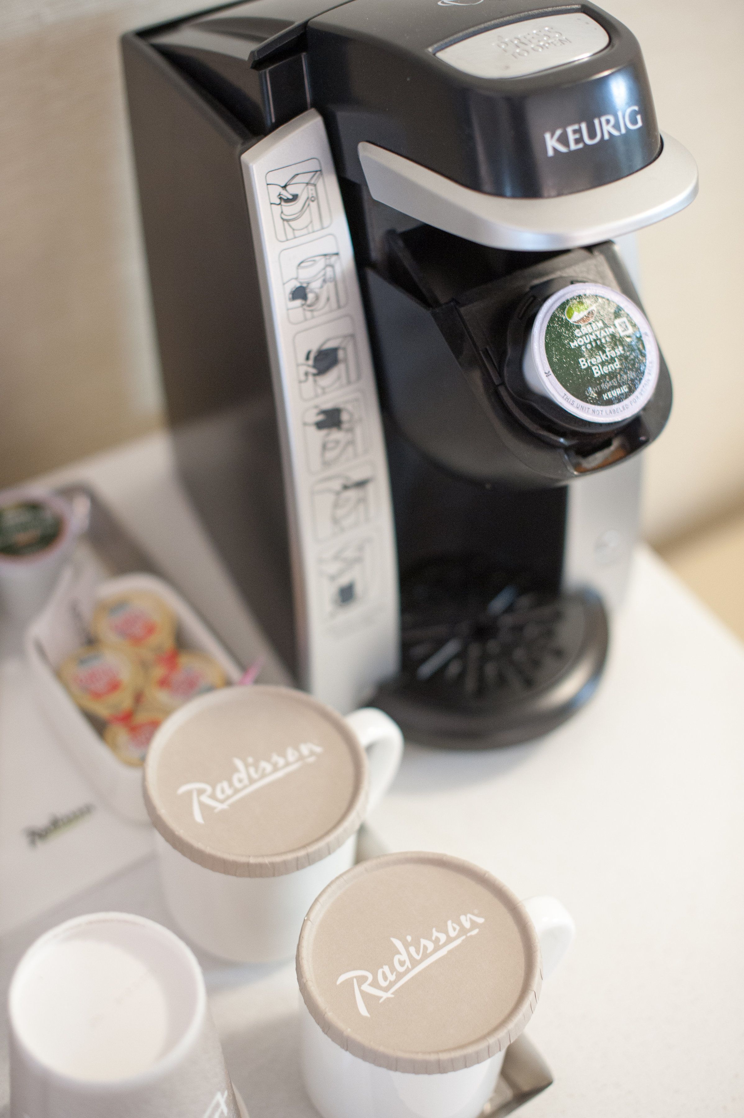 Eine Kaffeemaschine von Keurig mit Tropfschale, zwei weiße Tassen mit dem Schriftzug 'Radisson' und eine Joghurtportion.
