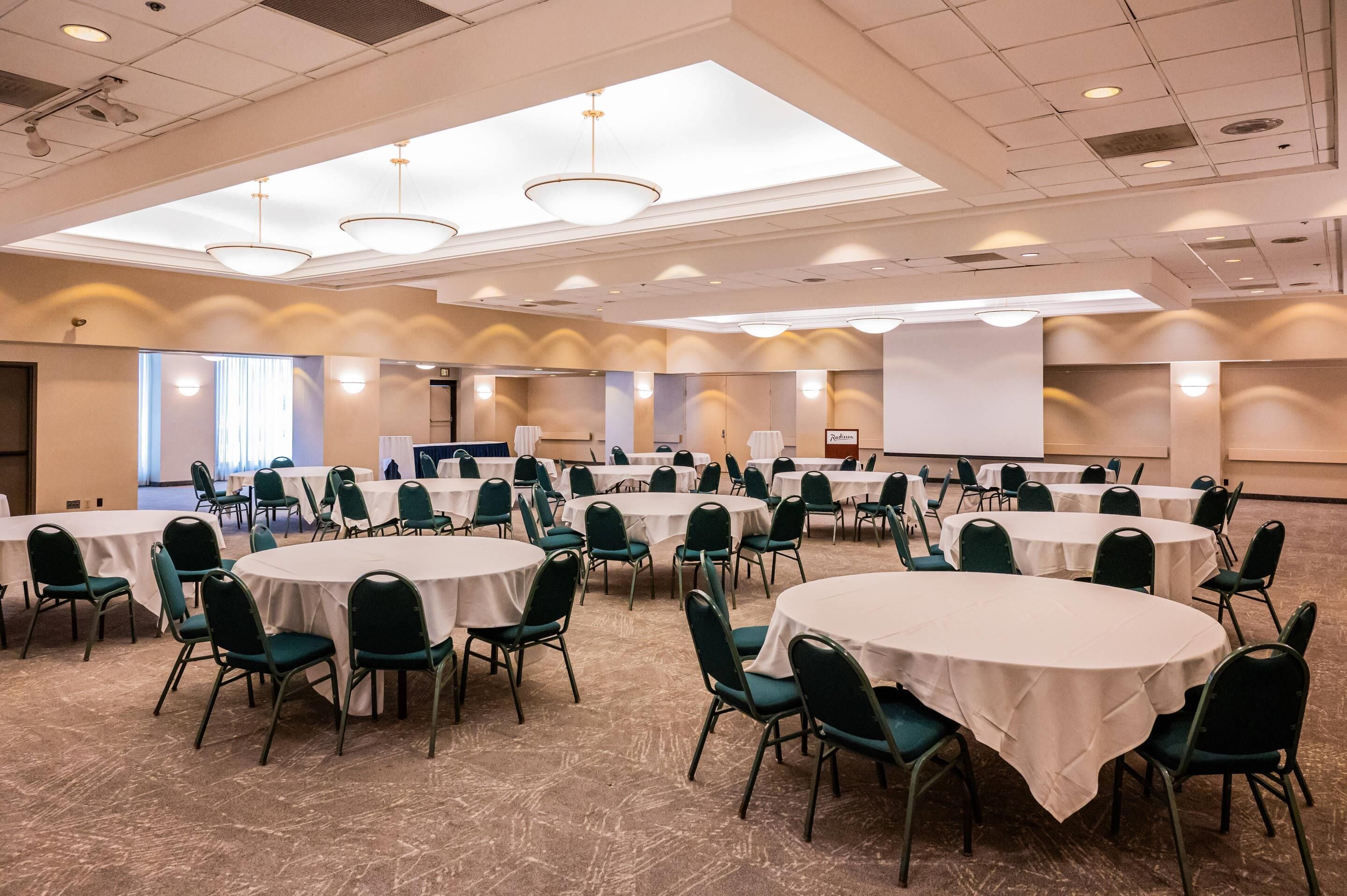 A conference room with multiple round tables and chairs, equipped with ceiling lights and a projection screen.