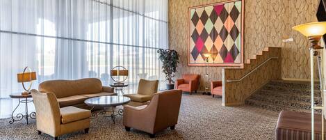Un hall de hotel con alfombra estampada, un ventanal con cortinas traslúcidas, un sofá, sillones, una mesita, una lámpara de pie y una escalera con revestimiento decorativo.