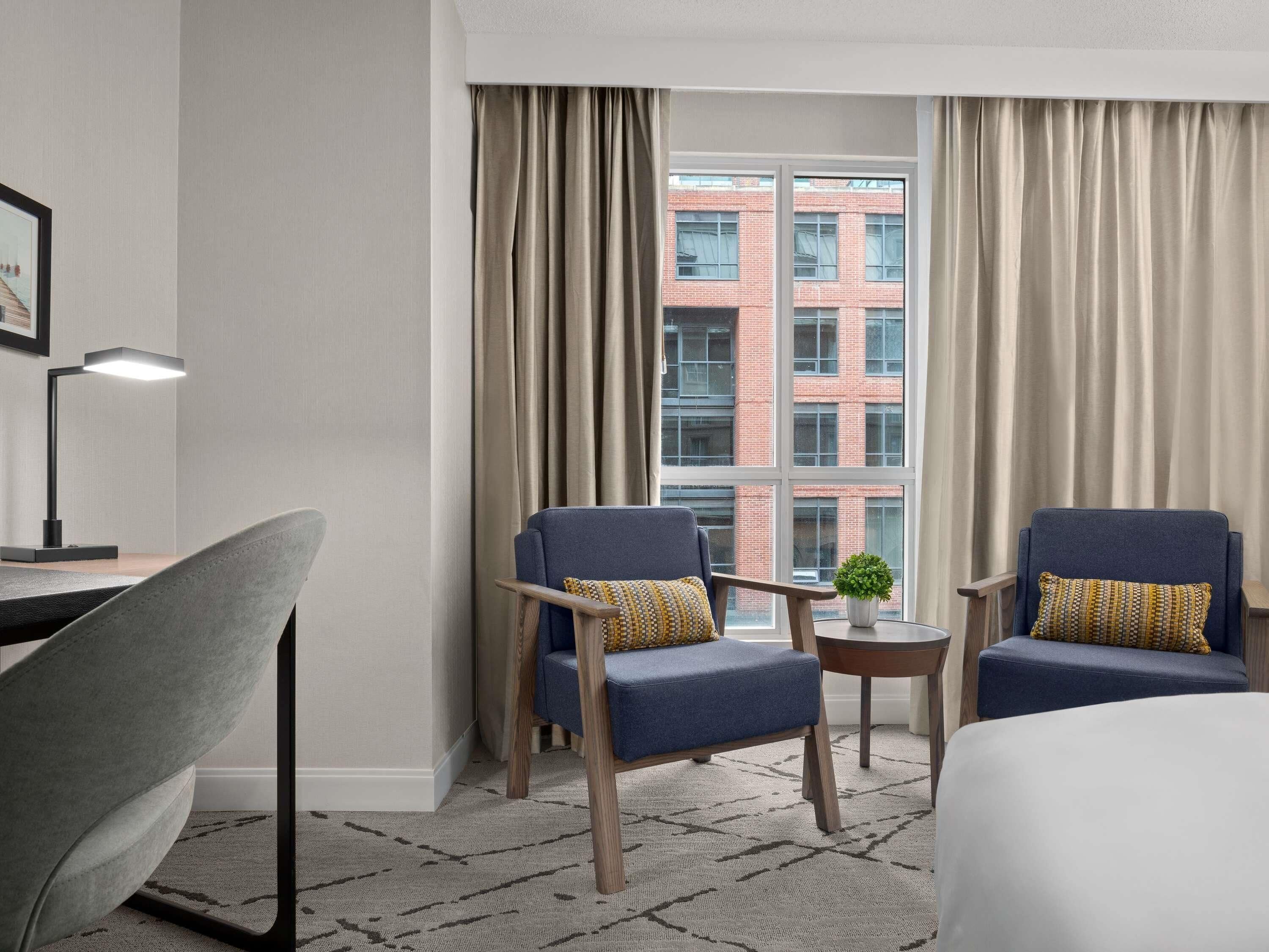 Quarto de hotel com duas poltronas, uma escrivaninha, uma luminária e vista para um edifício de tijolos por grandes janelas.