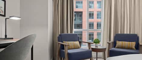 A hotel room with two armchairs, a desk, a lamp, and a view of a brick building through large windows.