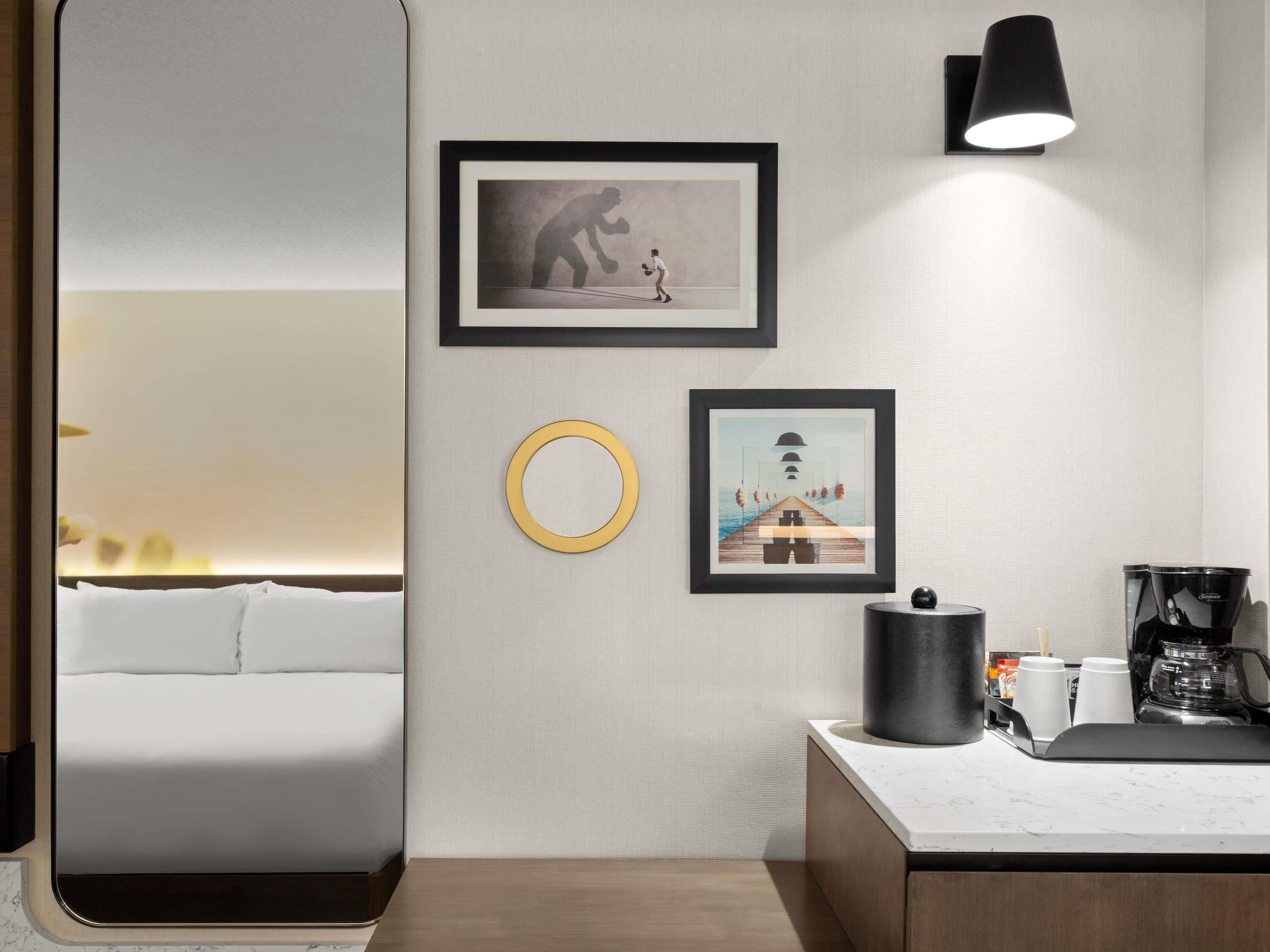 A hotel room with a bed, a desk with a coffee maker, and two framed artworks on the wall.
