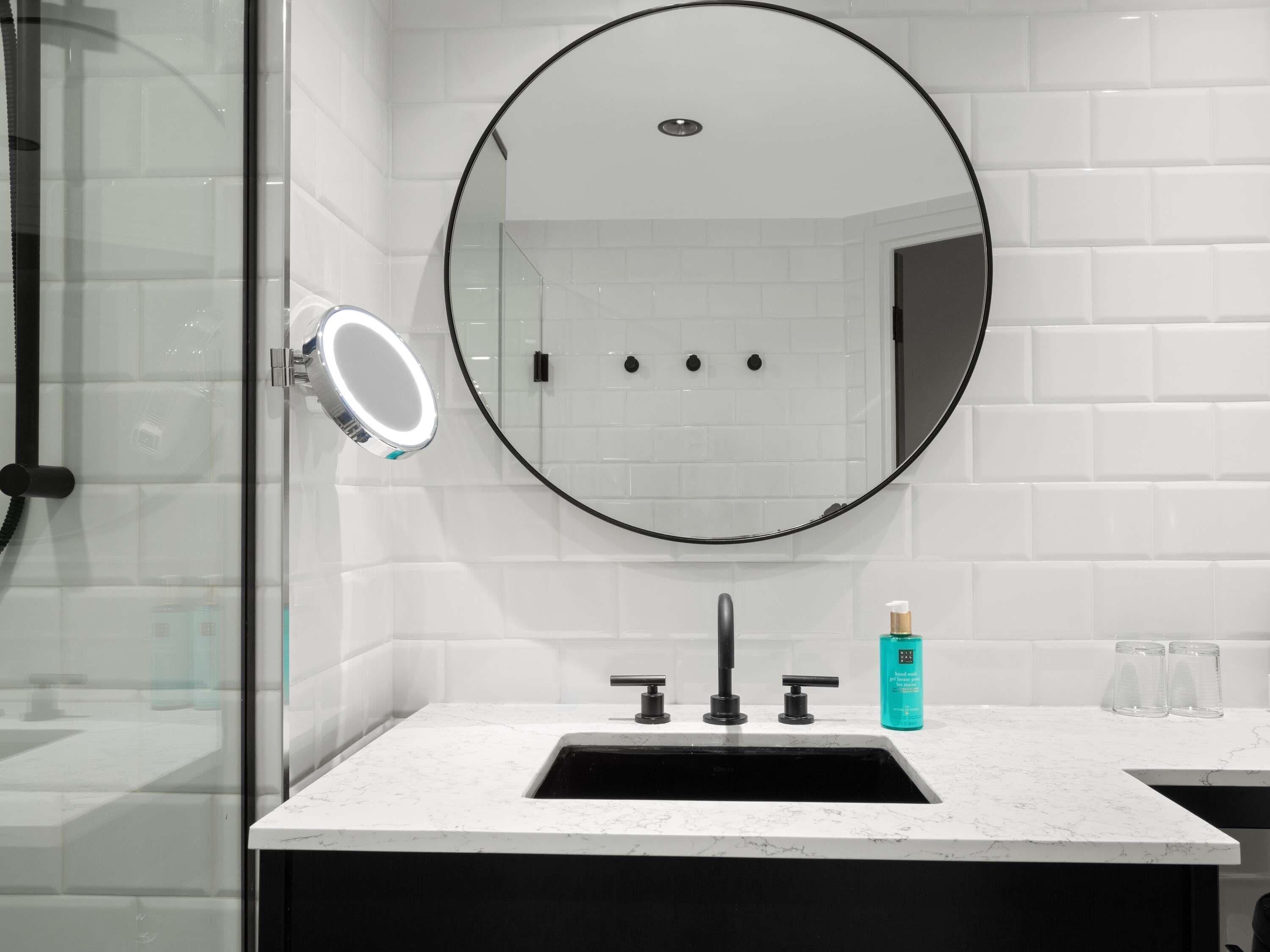 A bathroom with a round mirror, a sink with two faucets, a glass shower enclosure, and a white tiled wall.