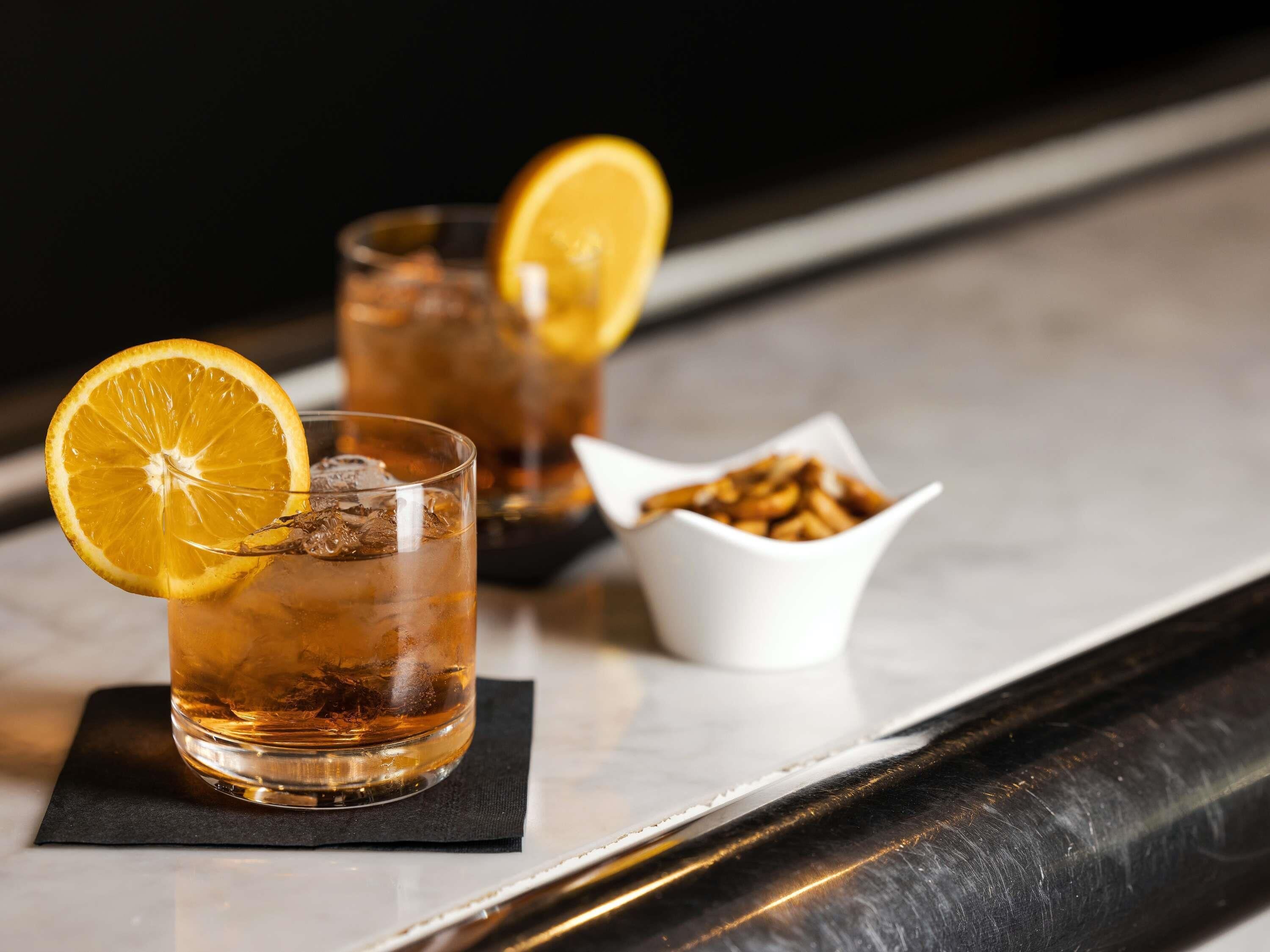 Two glasses of amber-colored beverage with ice and a slice of orange garnish, a small bowl of nuts, and a black napkin on a reflective surface.