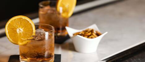 Two glasses of amber-colored beverage with ice and a slice of orange garnish, a small bowl of nuts, and a black napkin on a reflective surface.