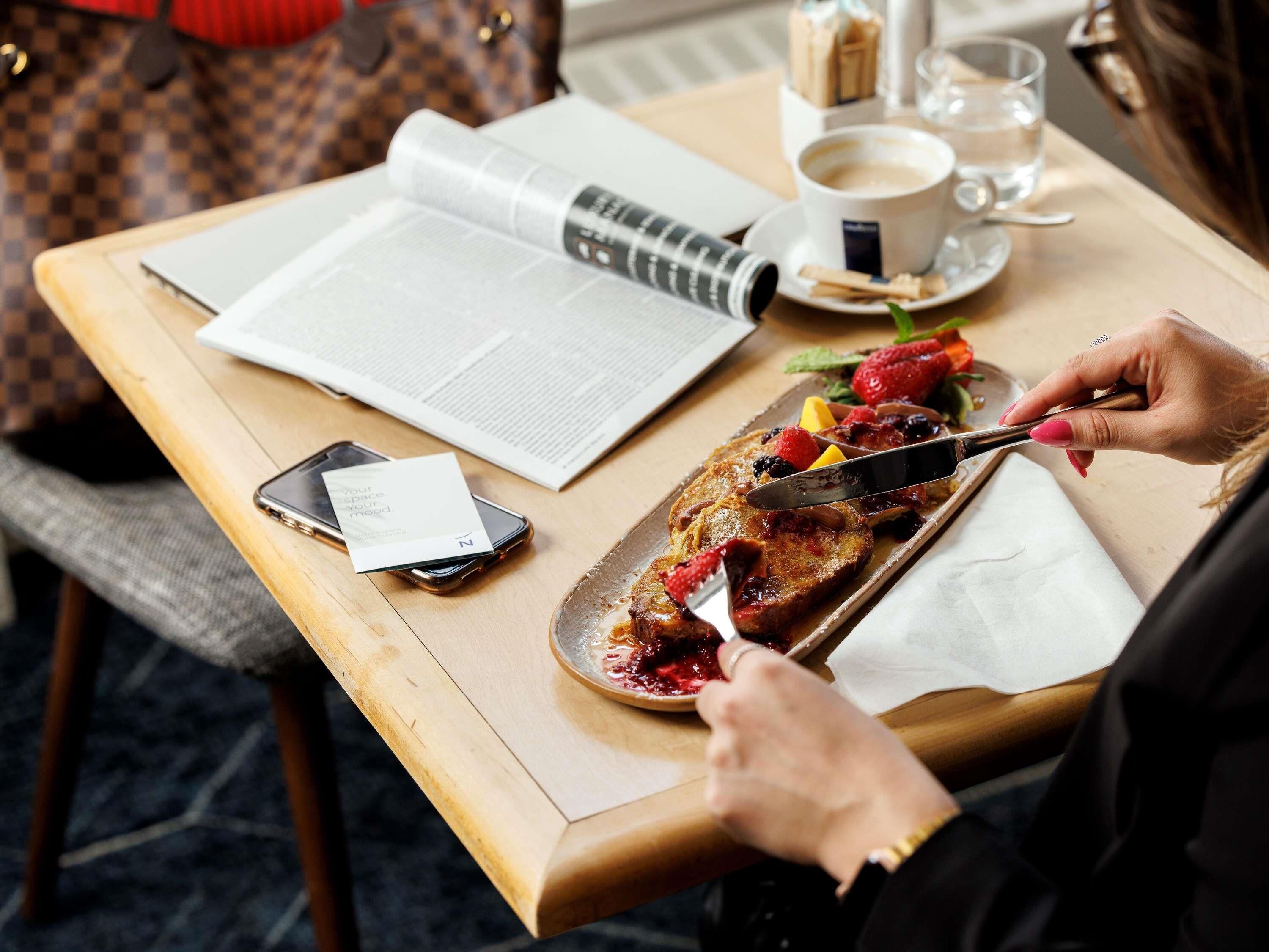 A person cutting a pancake with a knife, a cup of coffee, a book, and a smartphone on a wooden table.