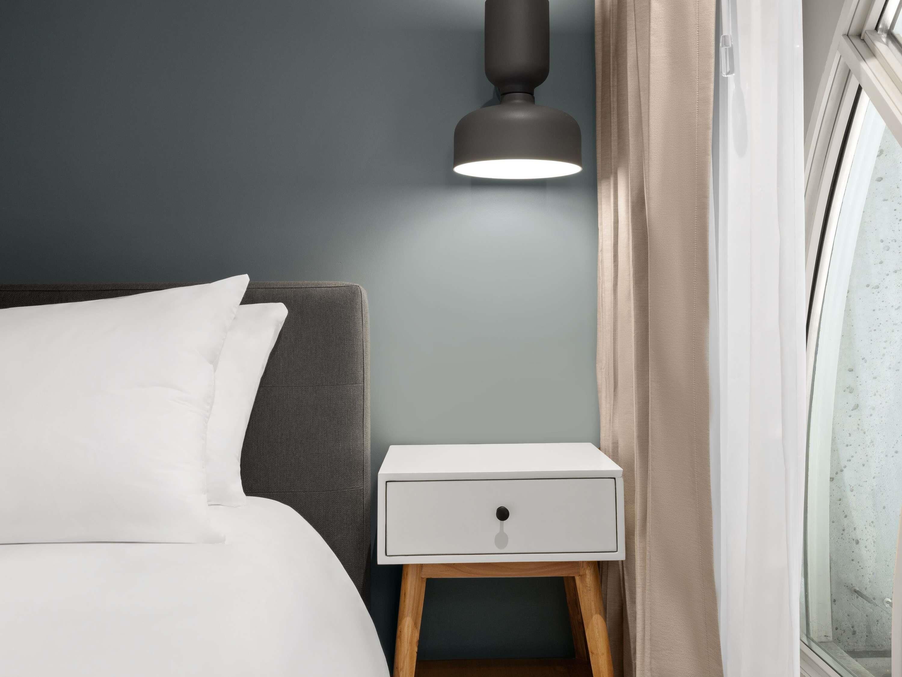 Ein ordentlich bezogenes Bett mit weißen Leinen, ein Nachttisch mit Schublade, eine moderne Lampe und ein Fenster mit Vorhängen.