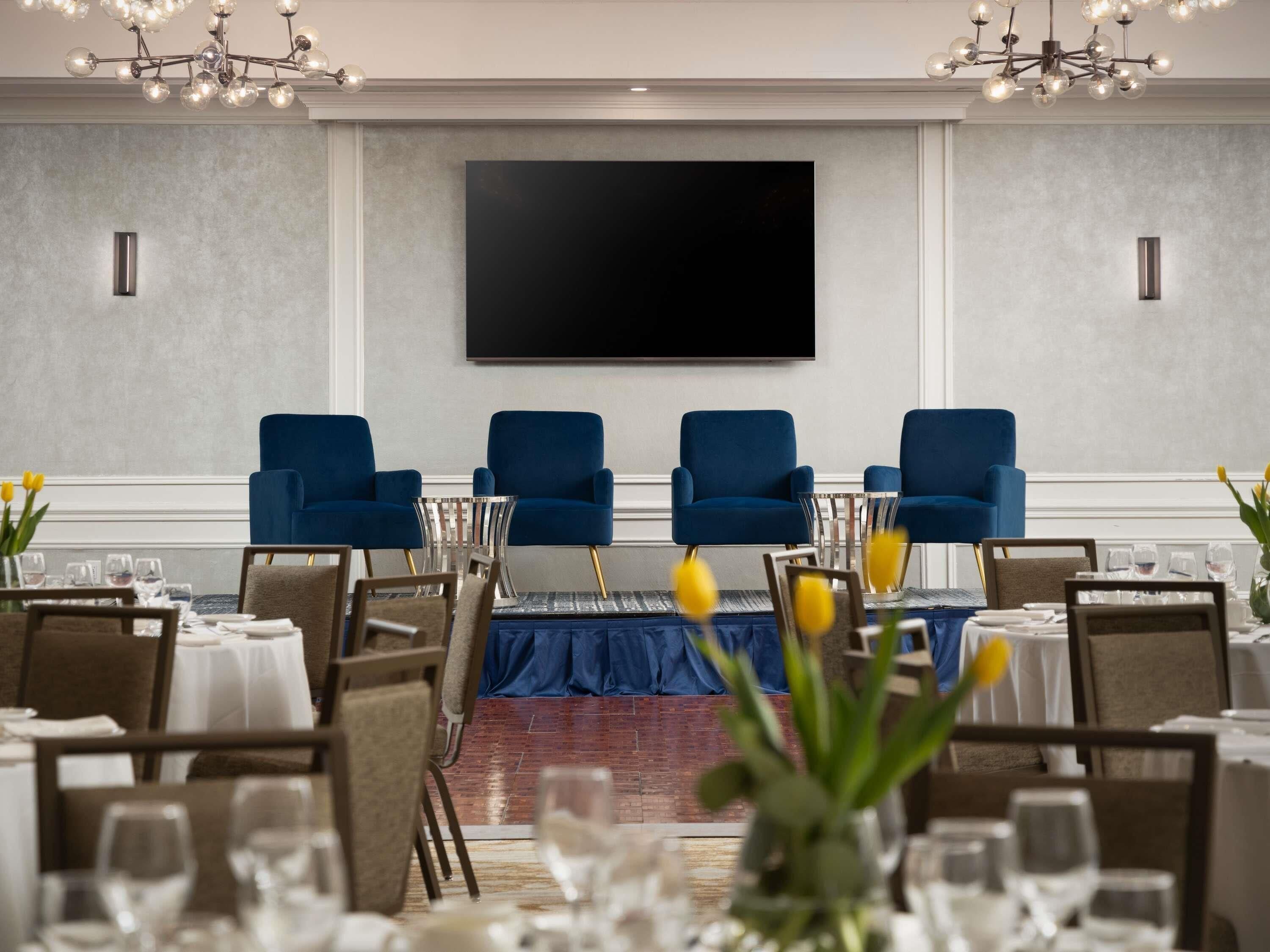 Un salón de conferencias con una televisión de pantalla plana grande, sillas azules y mesas dispuestas para una reunión.