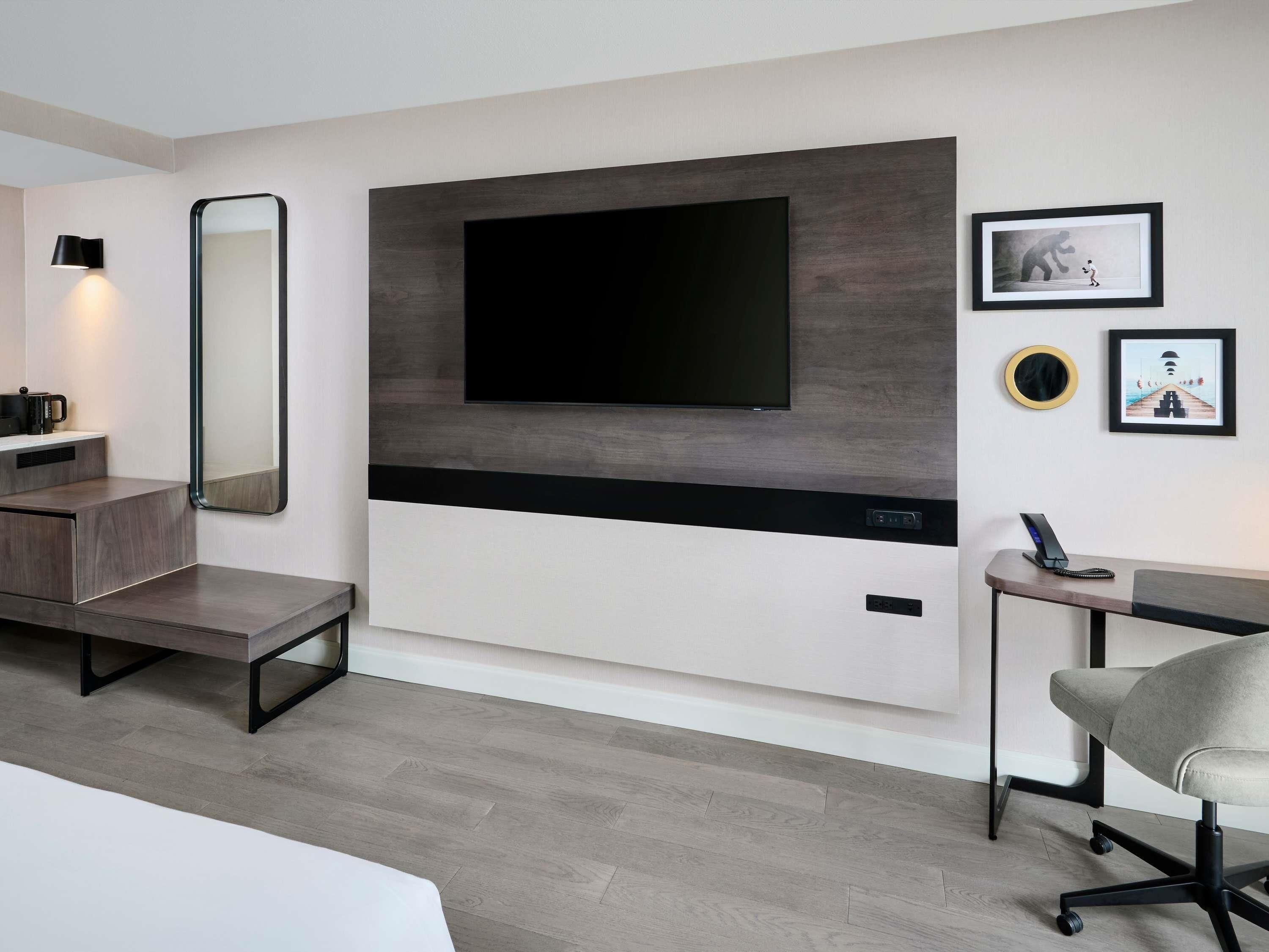 Et moderne hotelværelse med et stort fladskærms-TV monteret på væggen, et natbord med en lampe, en stol og et lille skrivebord med en telefon.