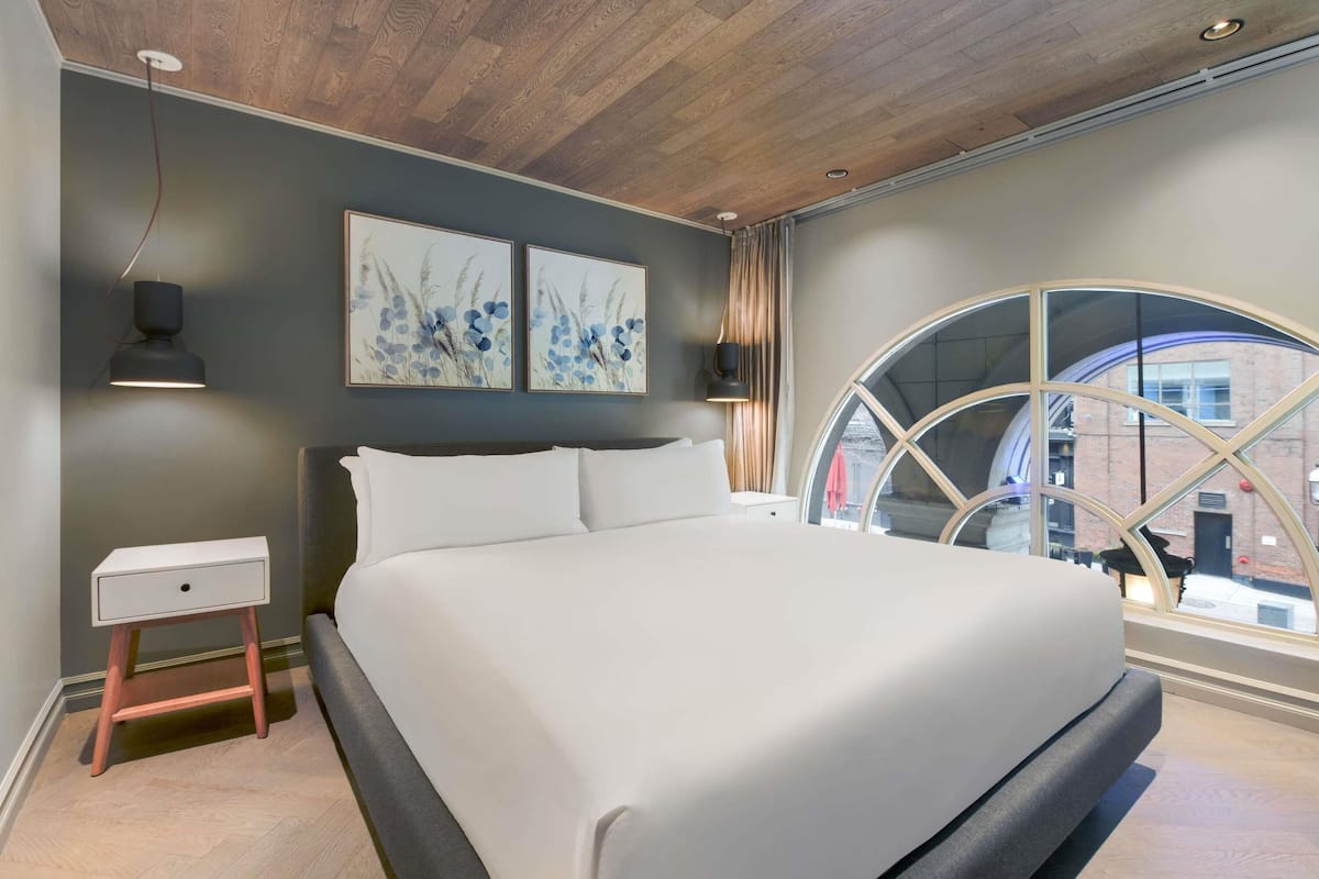 Un dormitorio moderno con una cama grande, una mesita de noche, una lámpara de pared y una ventana con vistas a la ciudad.