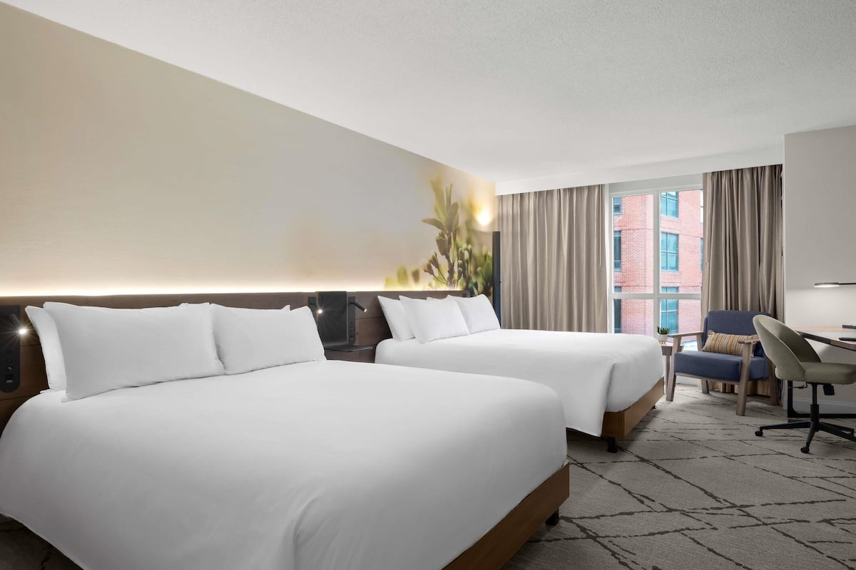 Habitación de hotel con dos camas, un escritorio, una silla y un ventanal con cortinas.