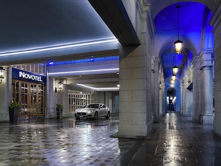 A hotel entrance with a car parked in front, a sign reading 'NOVOTEL,' and a wet floor indicating recent rain.