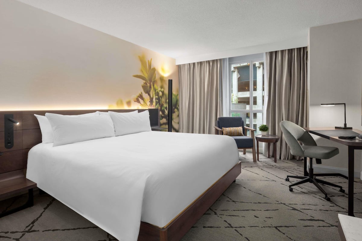 Habitación de hotel con una cama grande, un escritorio con silla, una mesita con lámpara y vistas al exterior a través de una ventana.