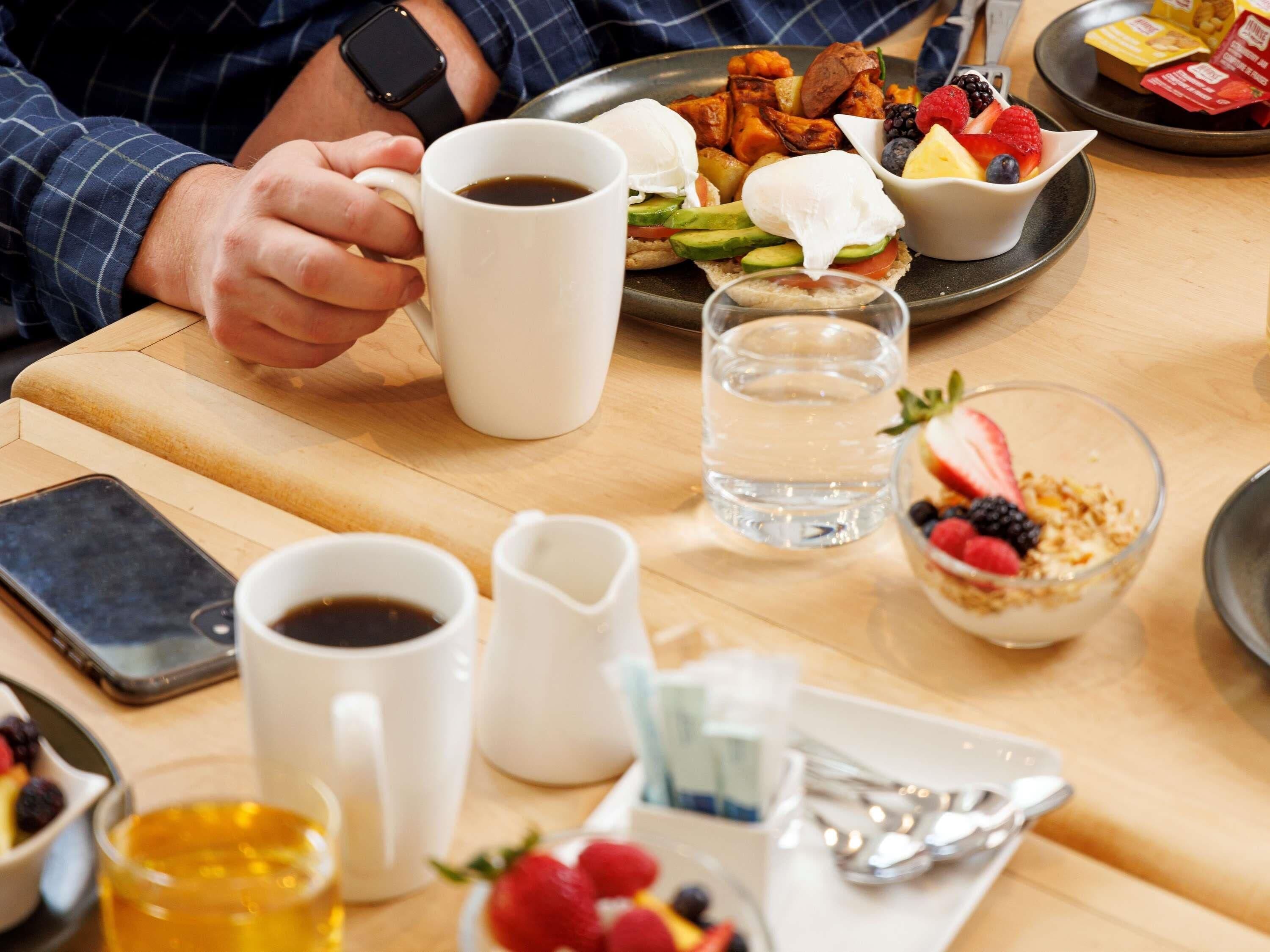 A breakfast setting with a plate of food, a coffee mug, a glass of water, a bowl of fruit, and a smartphone on the table.