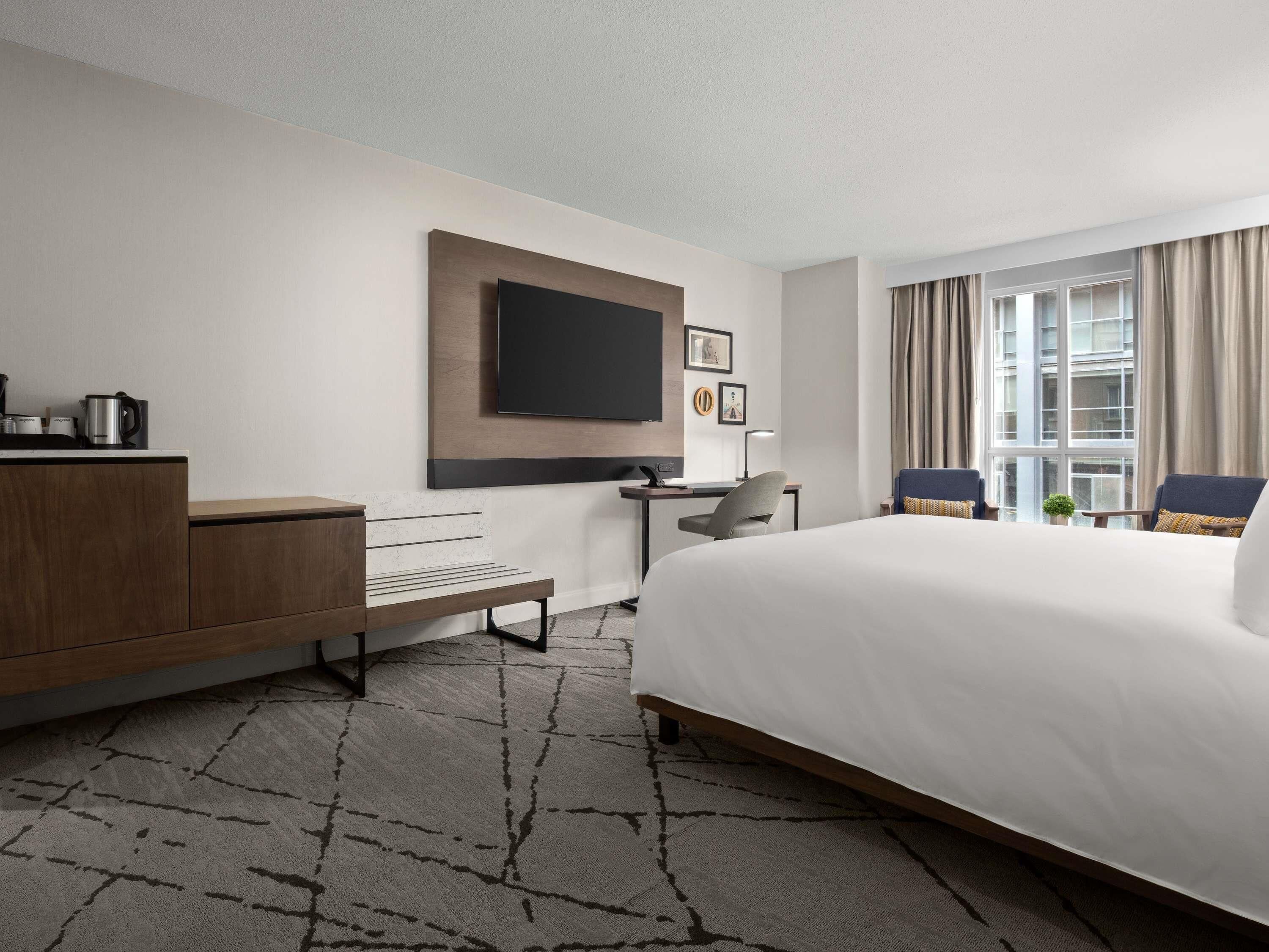 A hotel room with a large bed, a desk, a flat-screen TV mounted on the wall, and a window with curtains.