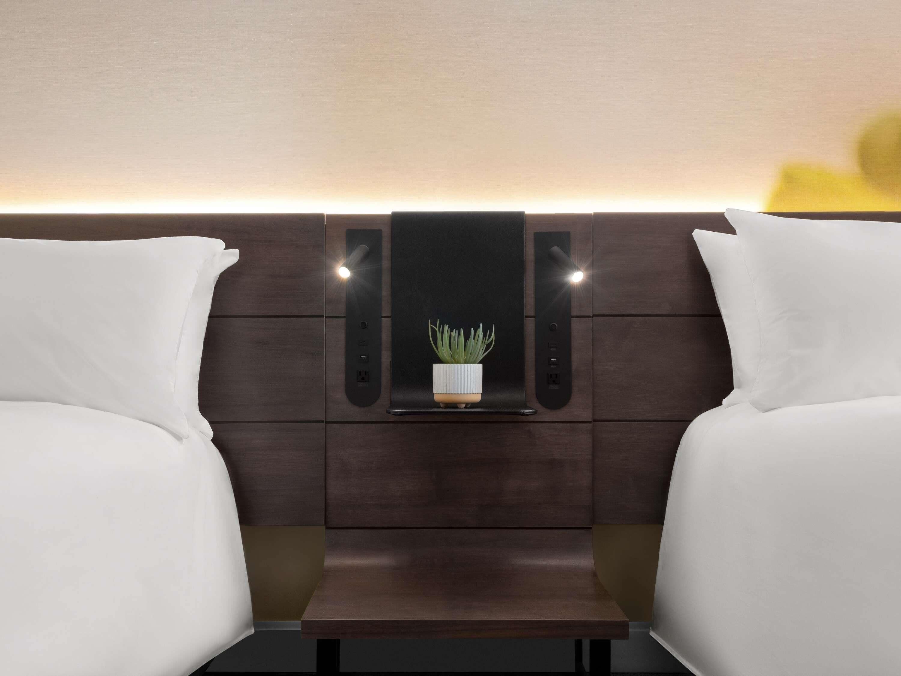 Une chambre d’hôtel avec une tête de lit en bois foncé, deux lits blancs, une petite table avec une plante et un luminaire fixé au mur.