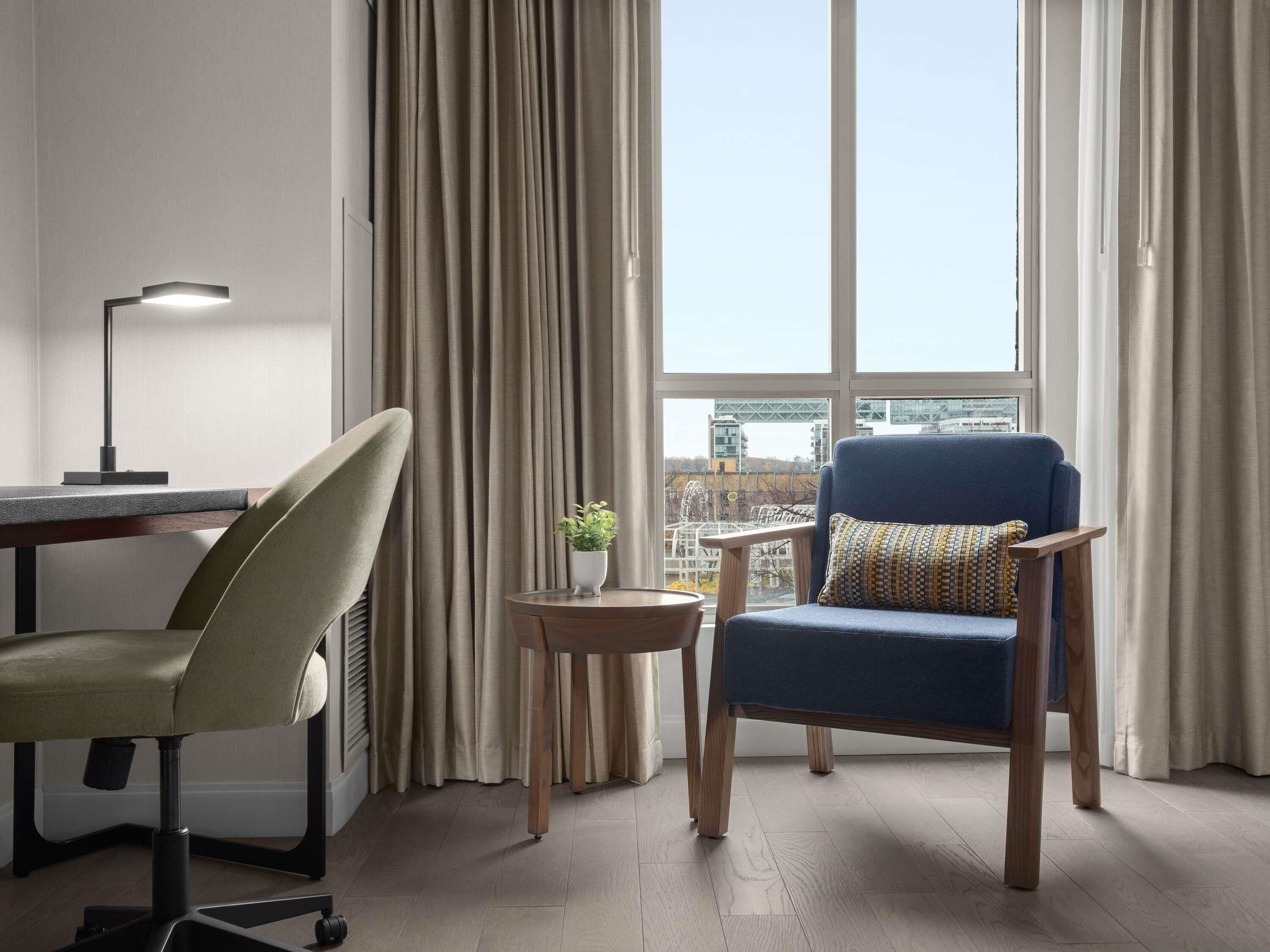 Una habitación moderna con escritorio, una silla, una mesita con una planta y vistas a edificios a través de amplios ventanales.