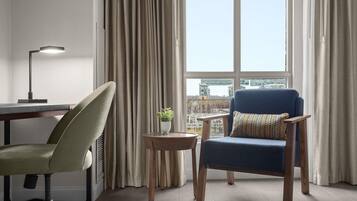 Una habitación moderna con escritorio, una silla, una mesita con una planta y vistas a edificios a través de amplios ventanales.