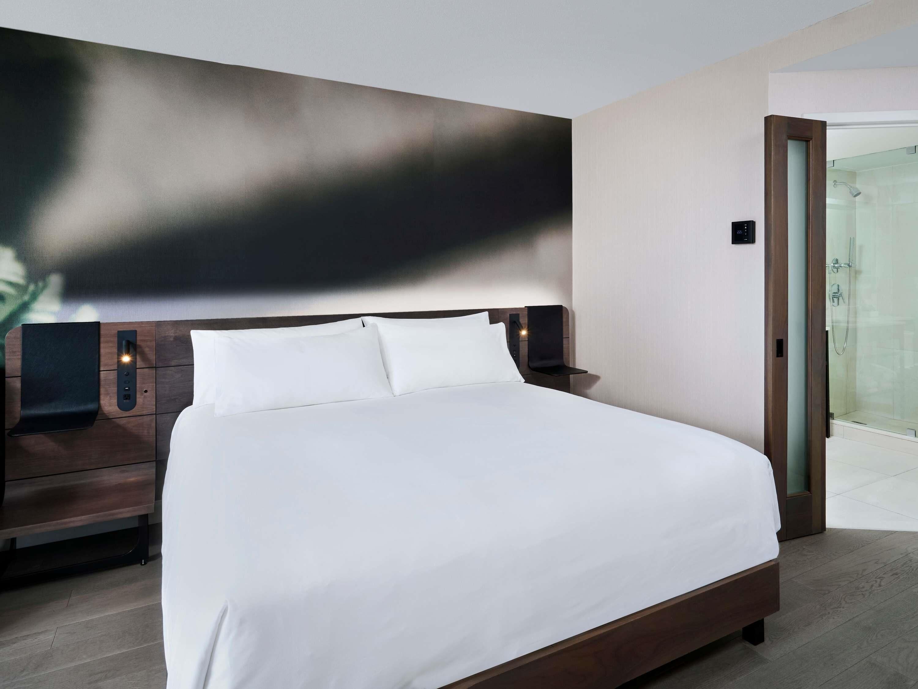 Ein modernes Hotelzimmer mit einem großen Bett, einem Nachttisch, einer wandmontierten Steuerung und einer Duschkabine aus Glas.
