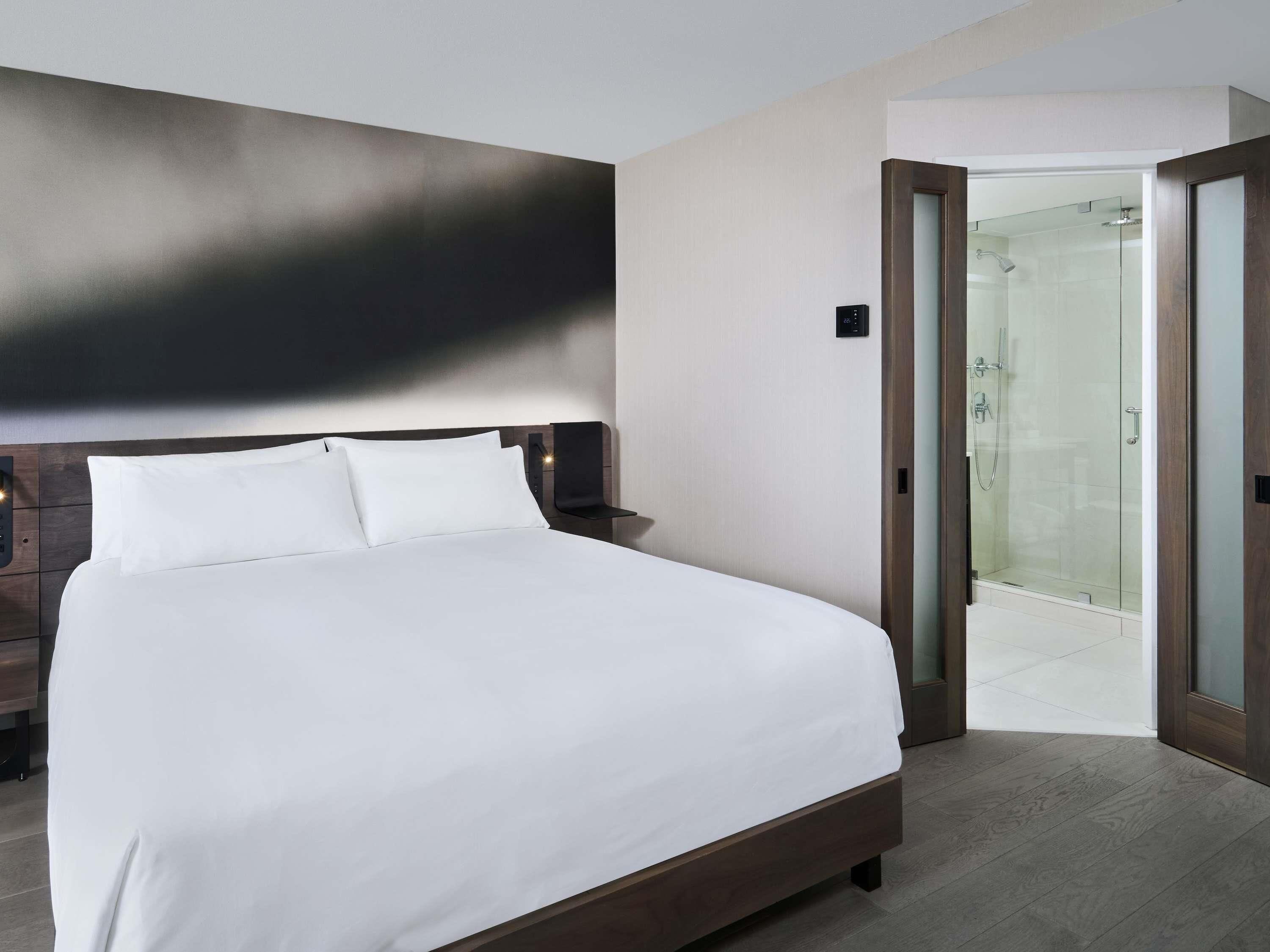 A modern hotel room with a large bed, a nightstand, a wall-mounted control panel, and a glass-enclosed shower area.