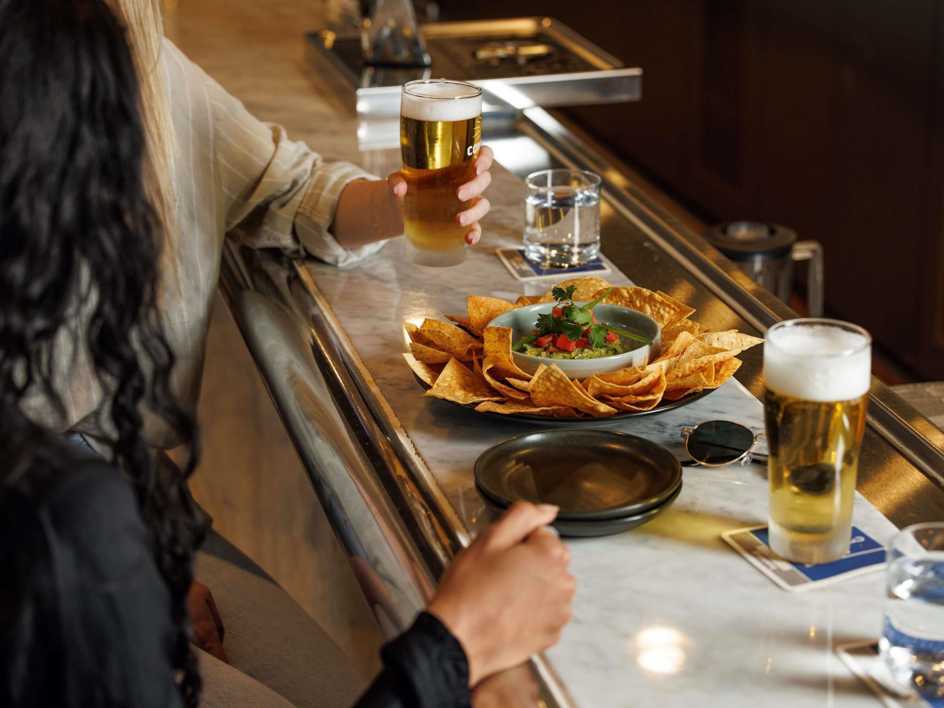A bar setting with a person holding a beer, a bowl of guacamole with chips, and a glass of water.
