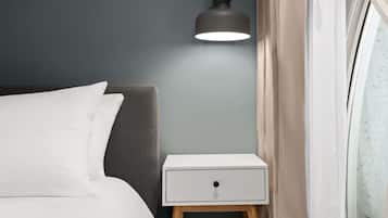 Un lit bien fait, avec du linge de lit blanc, une table de chevet dotée d’un tiroir, une lampe moderne et une fenêtre avec des rideaux.