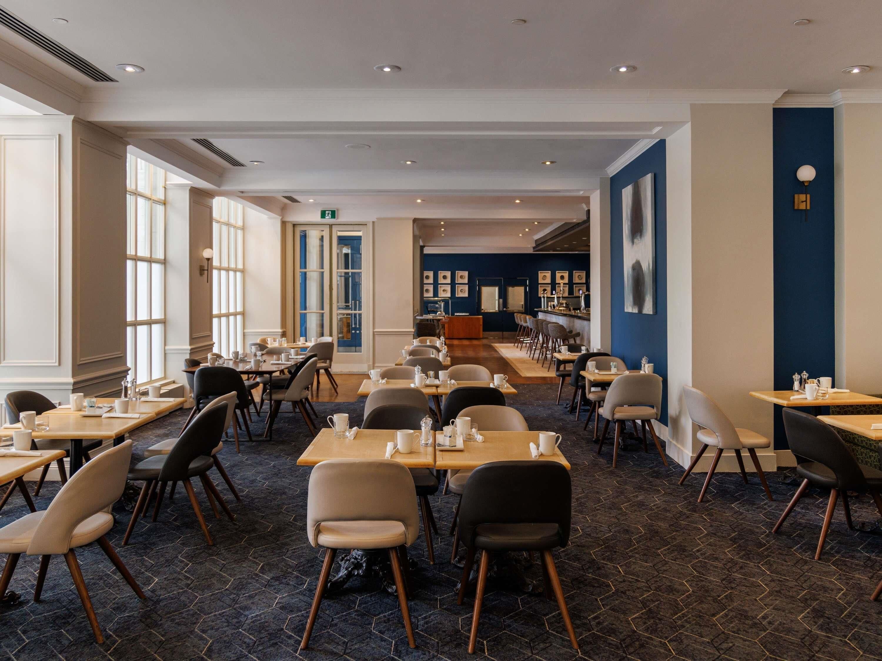 A spacious dining area with multiple tables and chairs, a bar in the background, and large windows allowing natural light.