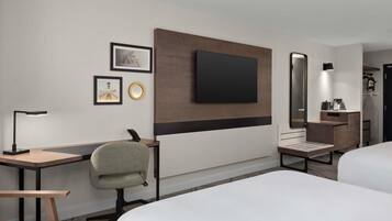 Habitación de hotel con dos camas, un escritorio con silla, televisor de pantalla plana en la pared, mesita de noche con lámpara y un espejo redondo pequeño.