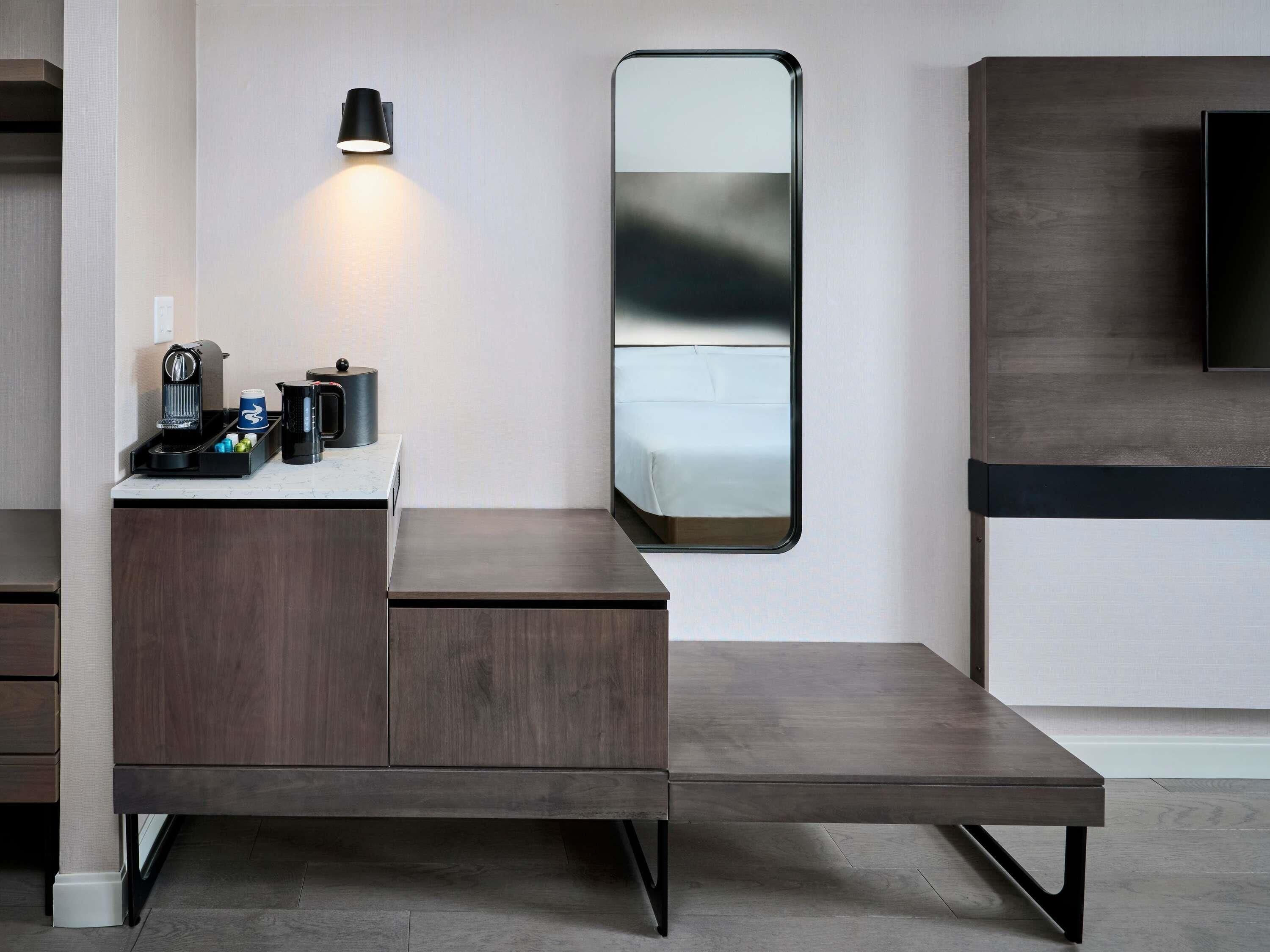 Una habitación de hotel moderna con un escritorio de madera, una cafetera, un espejo y un televisor.