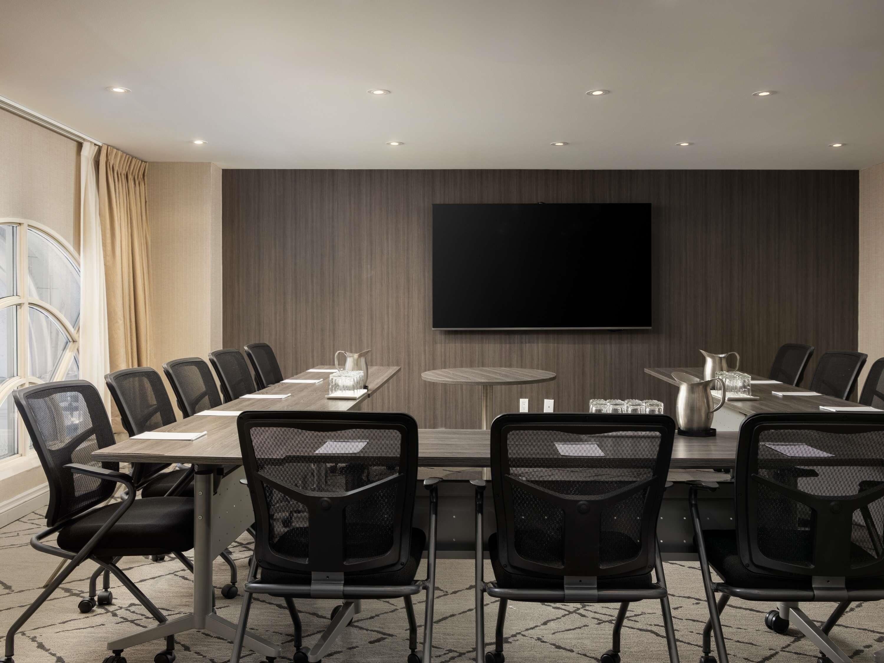 Une salle de conférence avec une longue table, des chaises, un écran de télévision et une petite table ronde sur laquelle se trouvent une carafe et des verres.