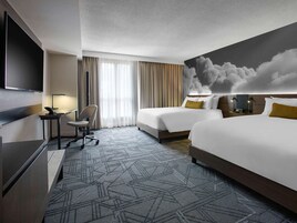 Quarto de hotel com duas camas, uma escrivaninha, uma cadeira, uma televisão e um grande mural com tema de nuvens na parede.