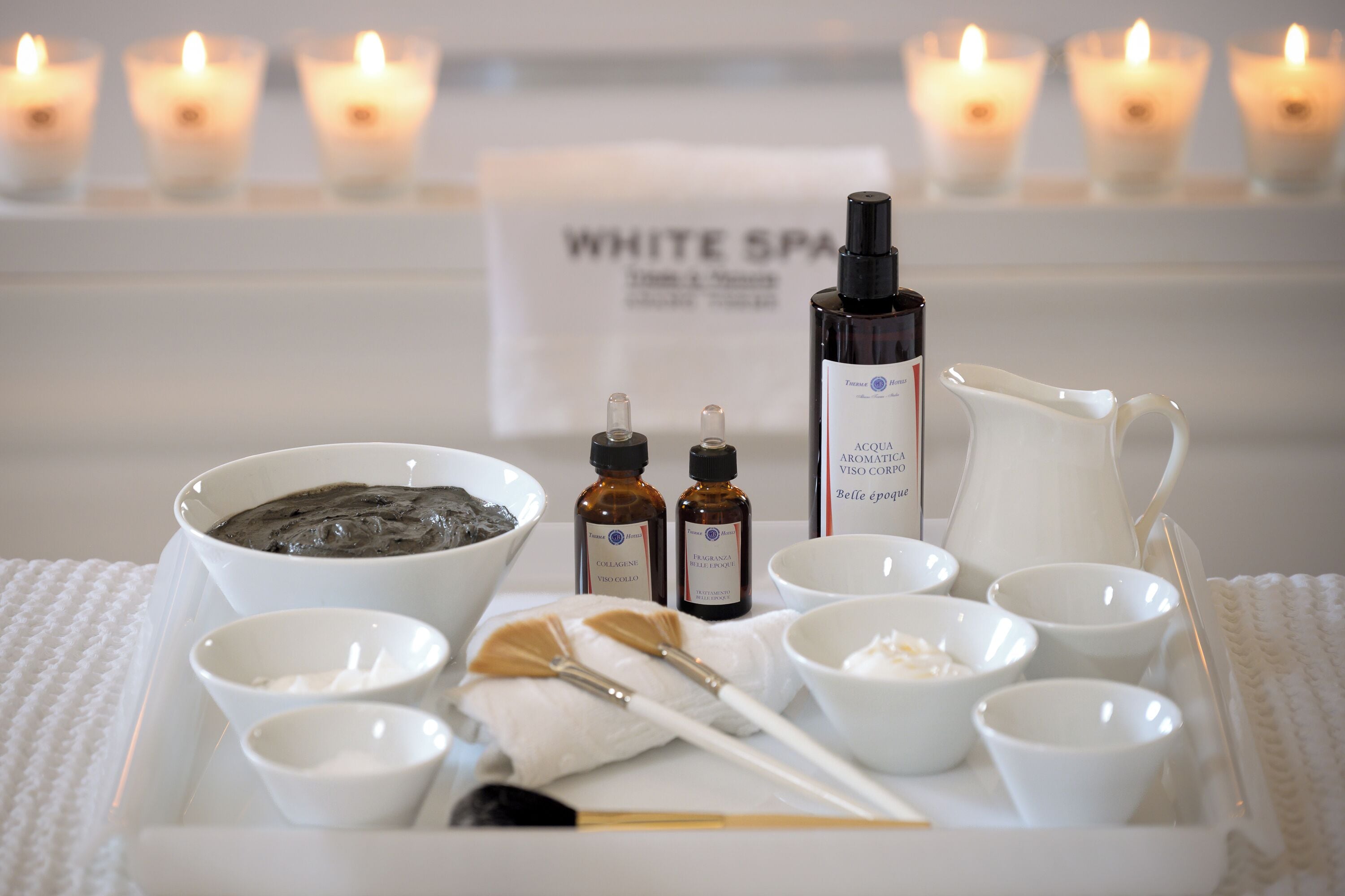 A tray with various spa products, including a bowl of black mud, two small bottles, a white pitcher, and several small bowls, set on a white towel with candles in the background.