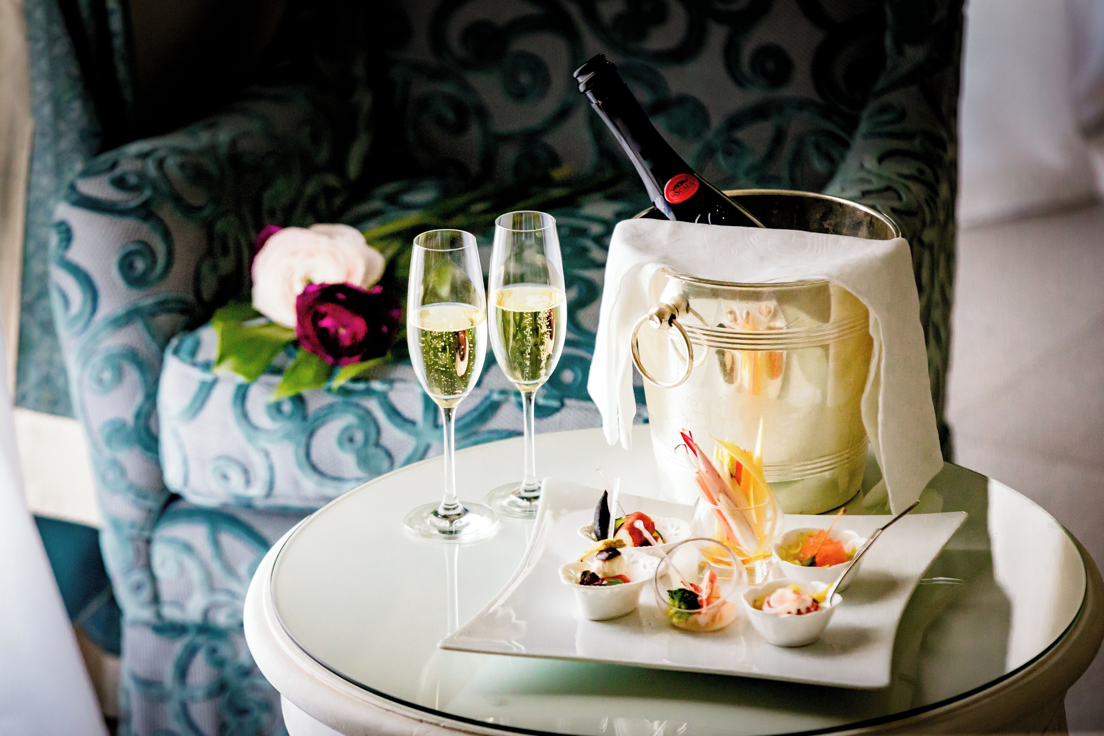 A table with two glasses of champagne, a bottle of champagne, and a pitcher with various appetizers.