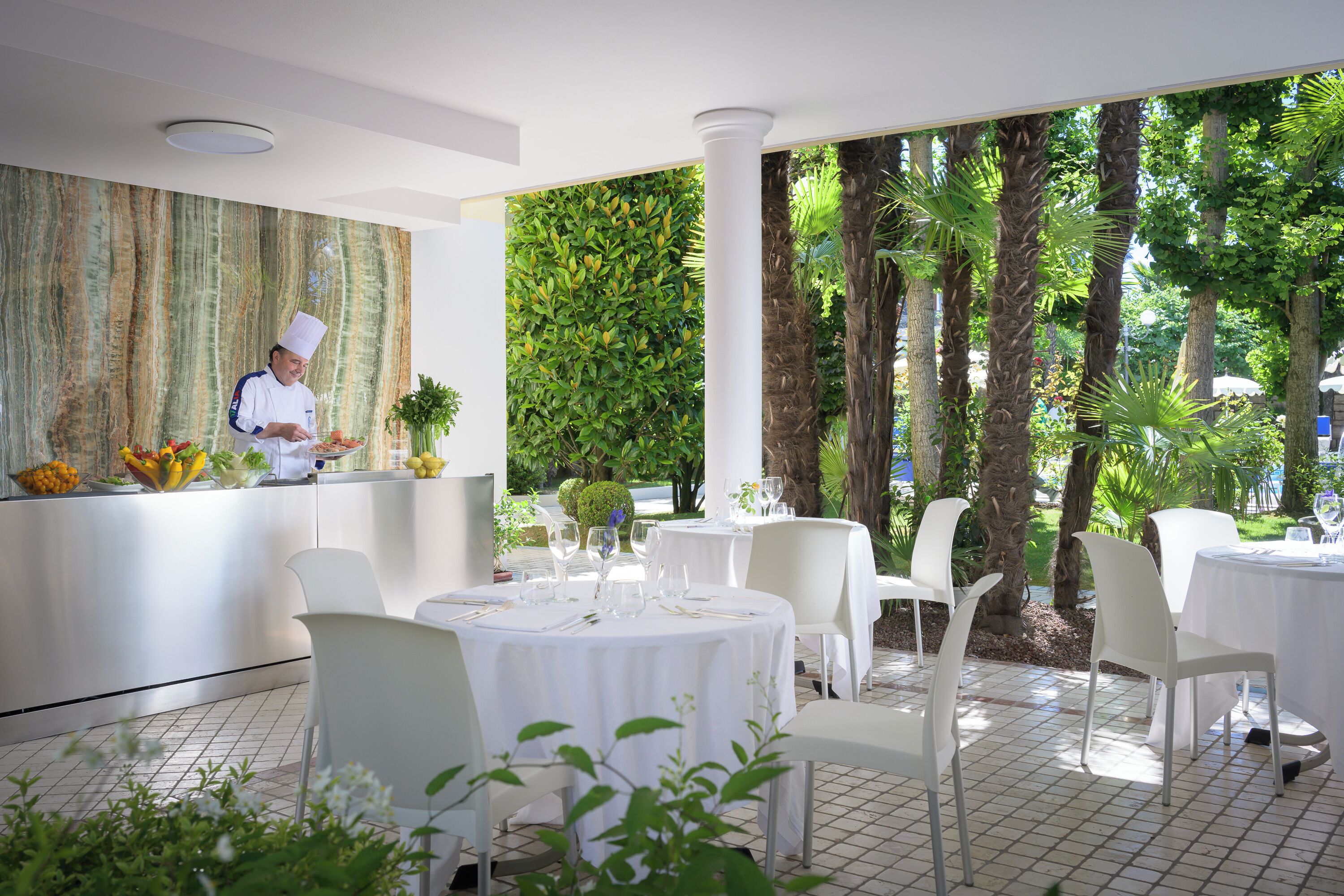 A restaurant with a chef preparing food, white tables and chairs, and a lush green garden view.
