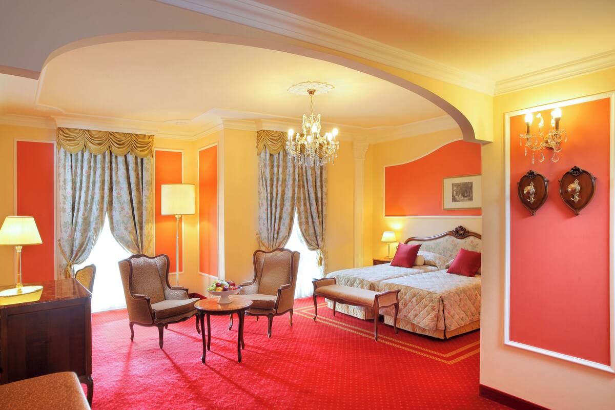 Une chambre d’hôtel avec un tapis rouge, un lit recouvert d’une couette rouge et blanche, deux fauteuils, une petite table sur laquelle se trouve un saladier de fruits, un lustre et un grand miroir au mur.