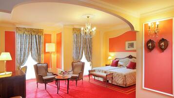 Une chambre d’hôtel avec un tapis rouge, un lit recouvert d’une couette rouge et blanche, deux fauteuils, une petite table sur laquelle se trouve un saladier de fruits, un lustre et un grand miroir au mur.