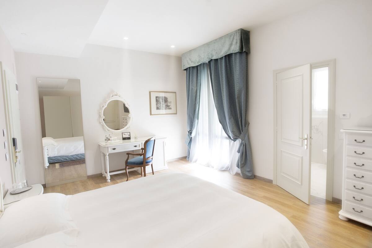 Une chambre à coucher avec un grand lit, une commode, un bureau avec une chaise, un miroir et une fenêtre avec des rideaux.