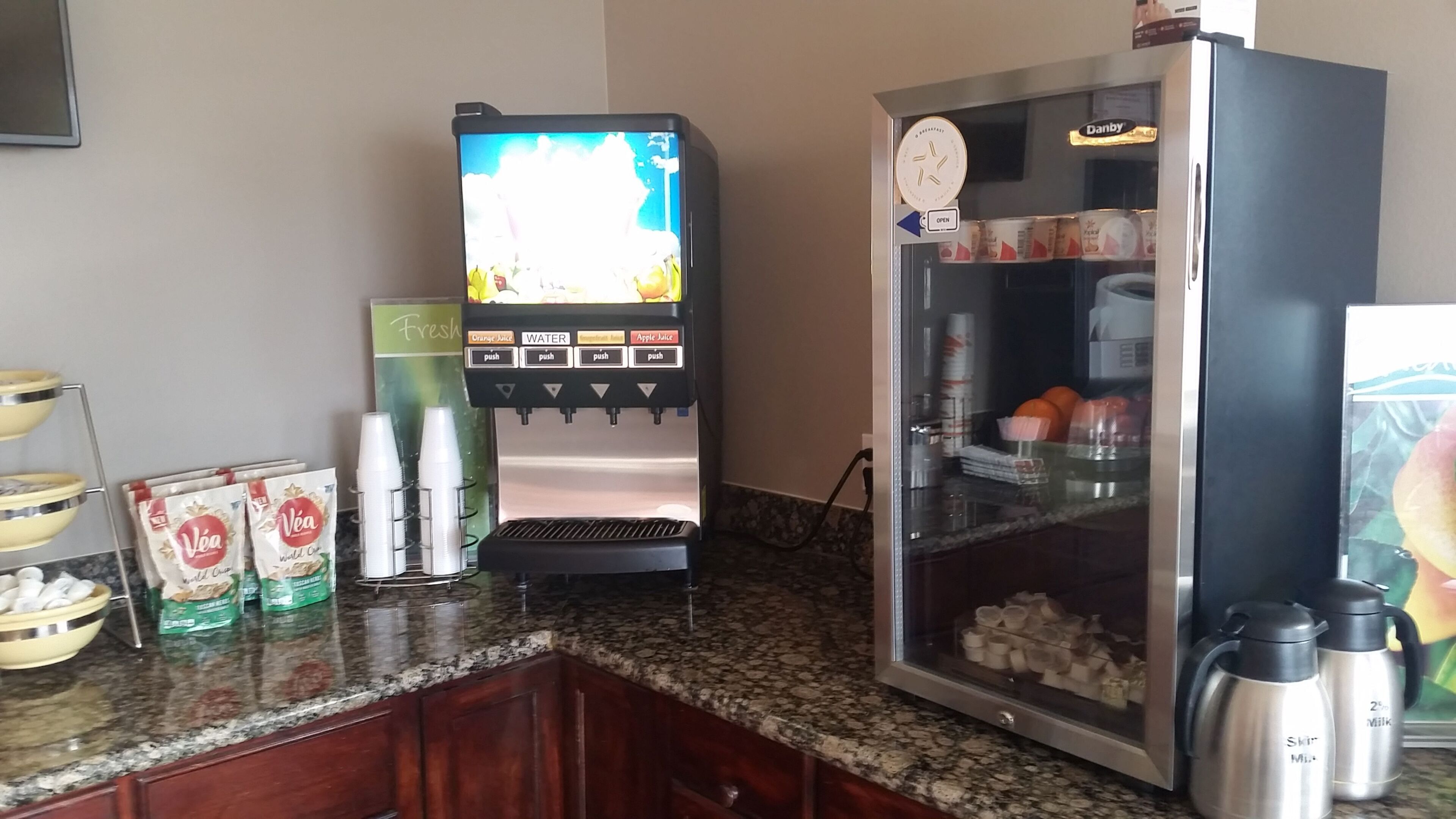 Un distributeur de boissons en libre-service proposant de l’eau, du jus de pomme et du jus d’orange, un réfrigérateur rempli de boissons et une machine à café.