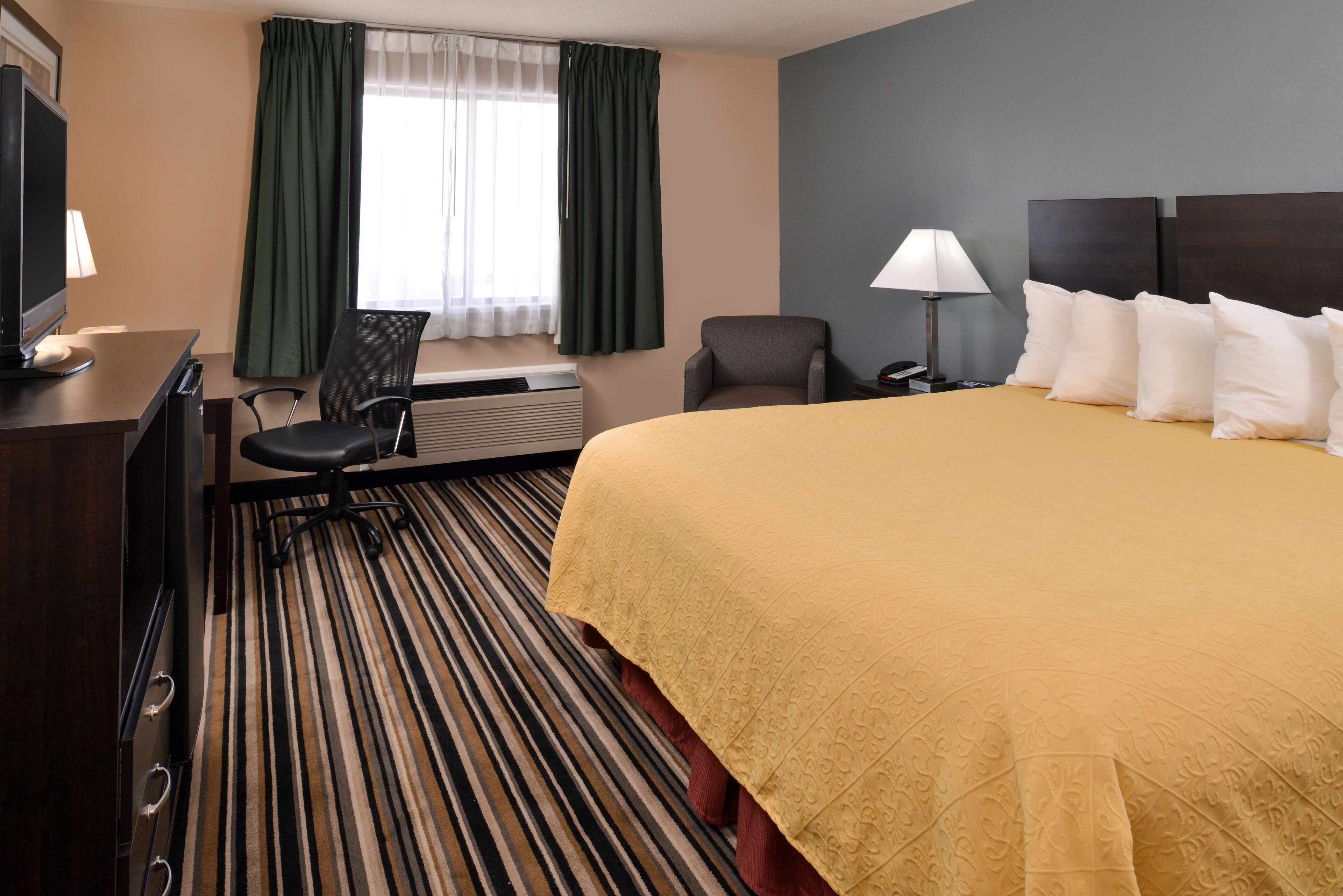 A hotel room with a bed, a desk with a computer, a chair, a lamp, and a window with curtains.