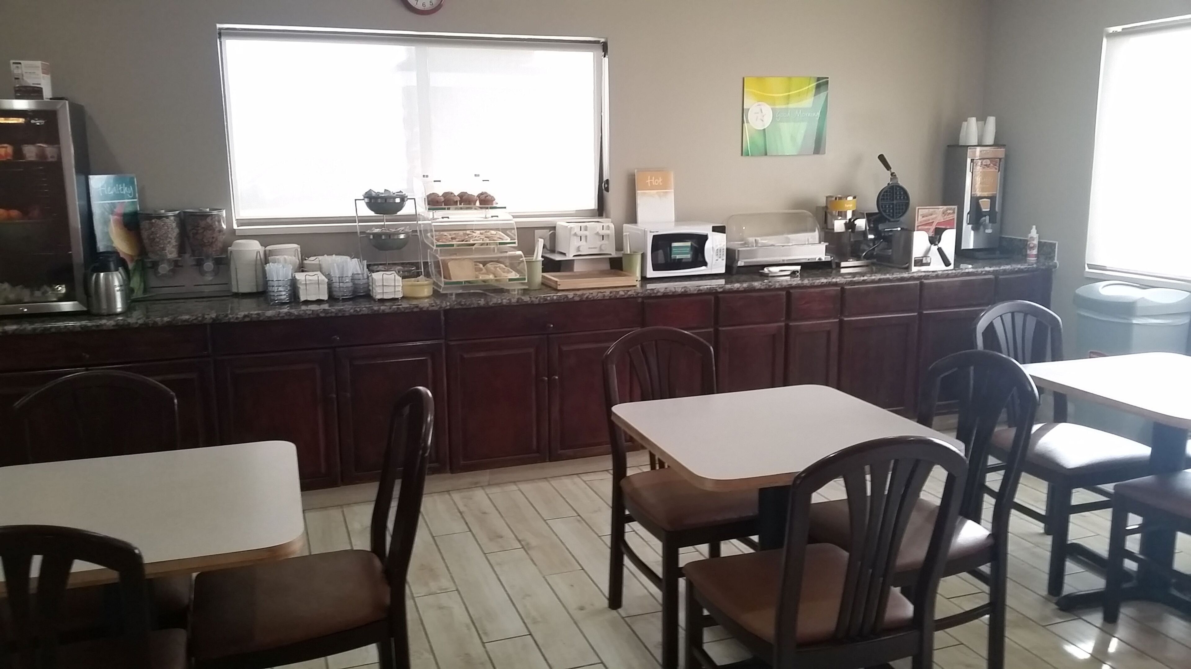 A kitchen with a large window, a microwave, a toaster, and various kitchen appliances. There are wooden cabinets and a dining area with chairs.
