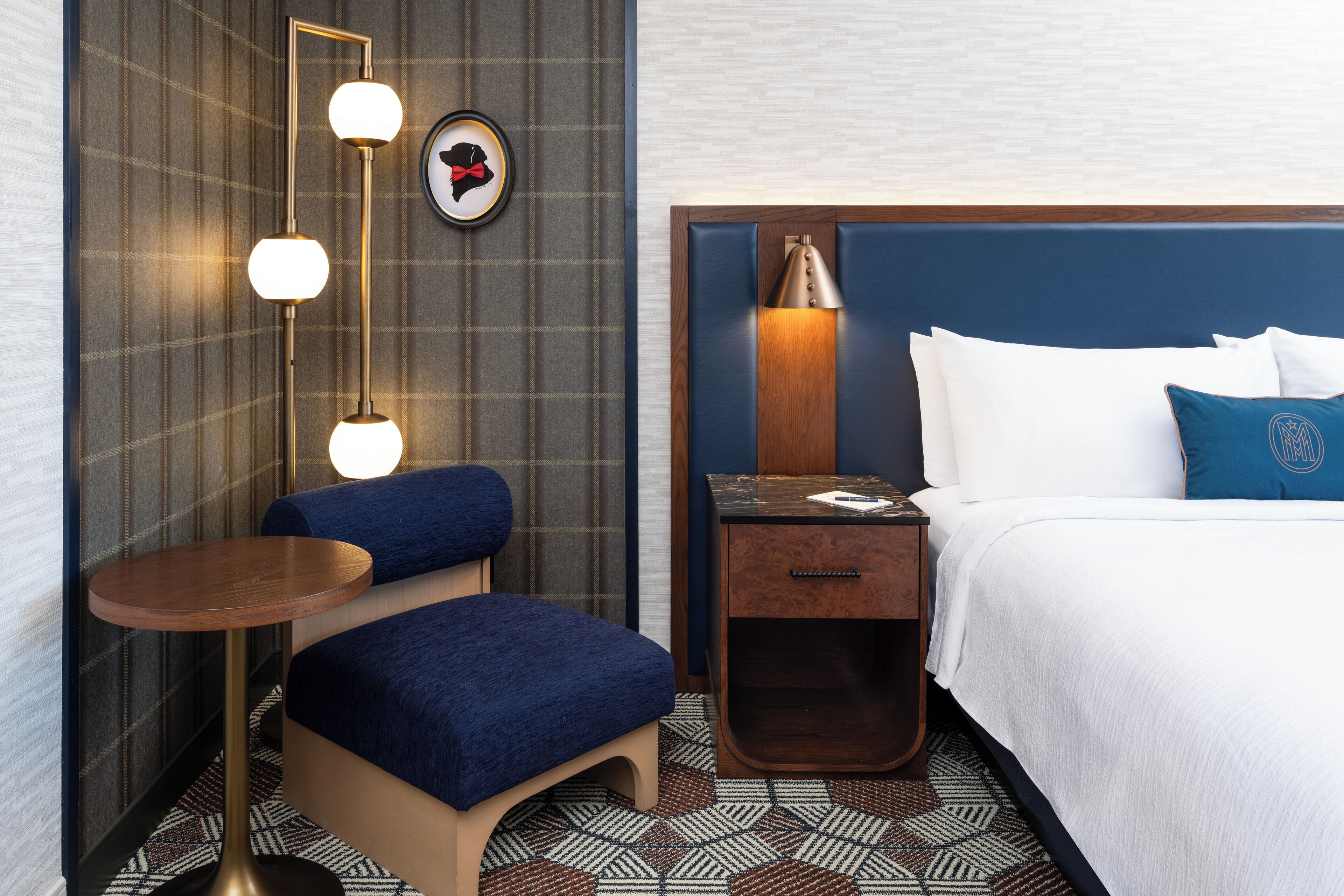 A hotel room with a bed, a nightstand, a desk, a chair, a lamp, and a wall-mounted light fixture.