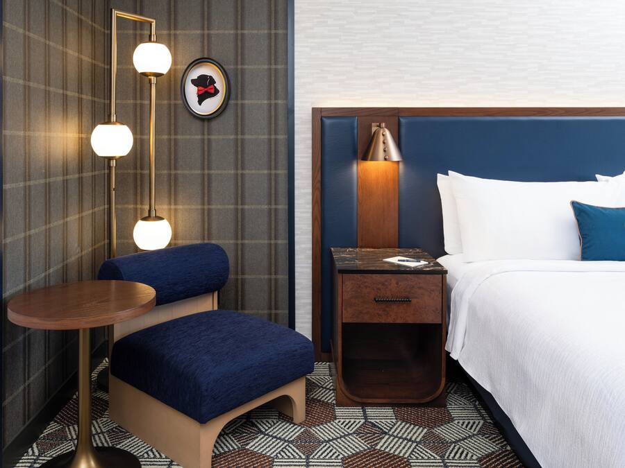 A hotel room with a bed, a nightstand, a desk, a chair, a lamp, and a wall-mounted light fixture.