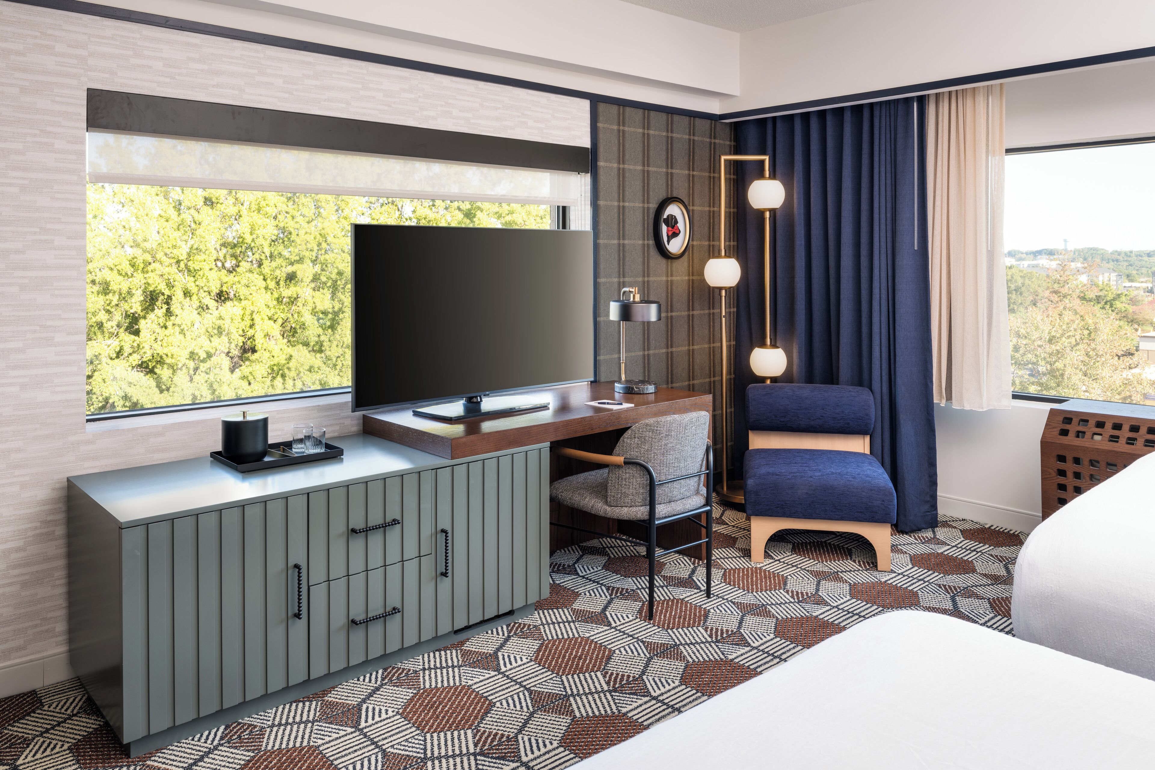 Une chambre d’hôtel moderne dotée d’une grande fenêtre, d’un téléviseur à écran plat, d’un bureau avec une lampe, d’une chaise bleue et d’un tapis à motifs.