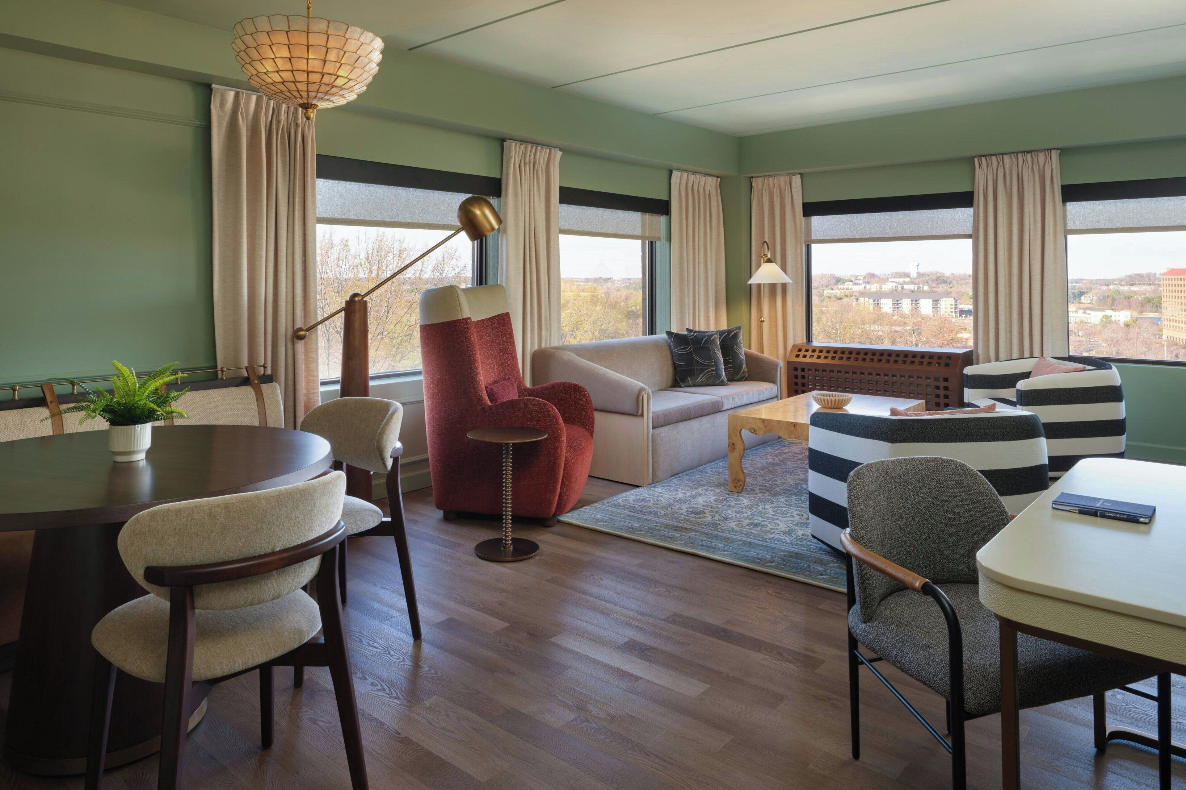 Una sala de estar moderna con una mesa redonda de comedor, un sillón rojo, un sofá y vistas de la ciudad a través de grandes ventanales.