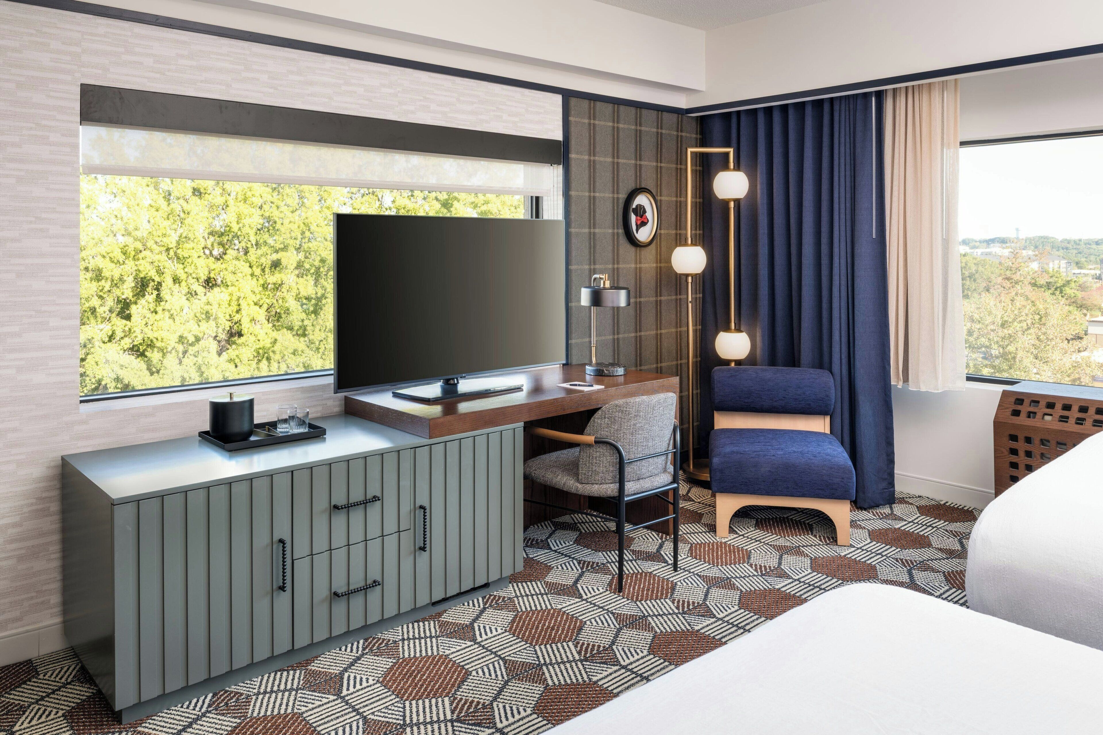A hotel room with a large window, a flat-screen TV, a desk with a chair, a sofa, and a patterned rug.