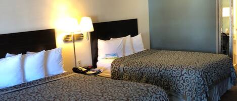 Habitación de hotel con dos camas, cada una con una colcha estampada y almohadas blancas. Hay una mesita de noche con una lámpara y un teléfono. La habitación tiene un diseño sencillo y limpio, con paredes azul claro y suelo alfombrado.