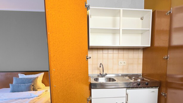 A compact hotel room with a kitchenette, a bed with a 'Feel at Home' sign, and a small bathroom. - Coffee Fellows Hotel Frankfurt-Langen (Langen)