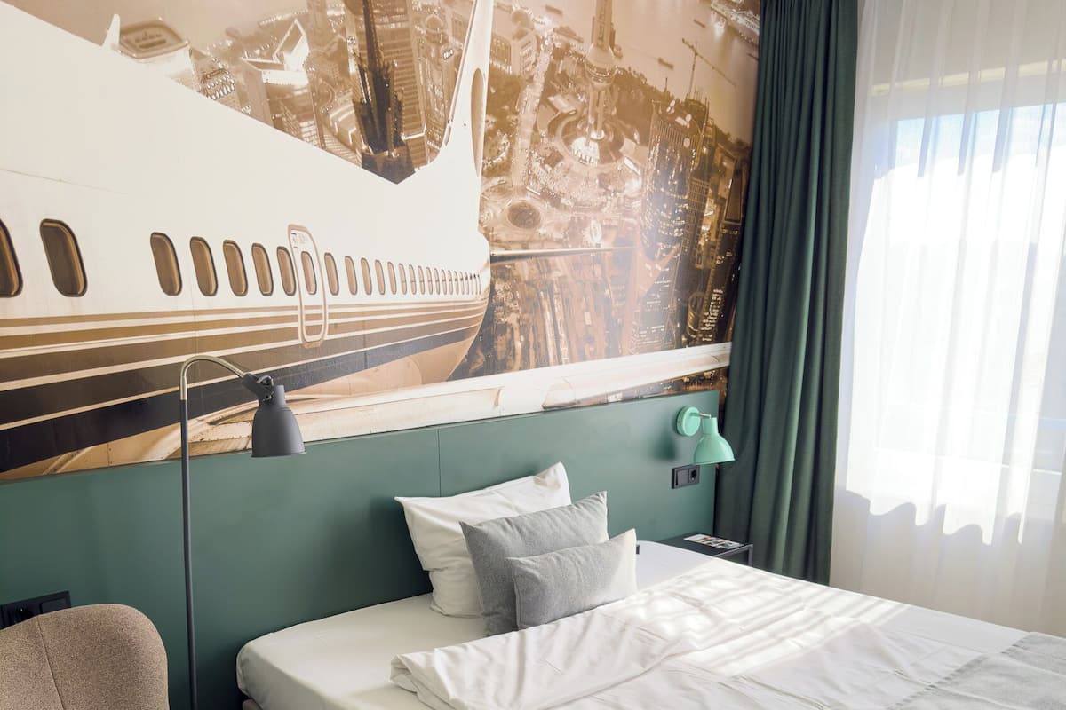 Ein Hotelzimmer mit Bett, Sessel, Stadtbild-Wandgemälde und einem fenster mit Vorhängen.