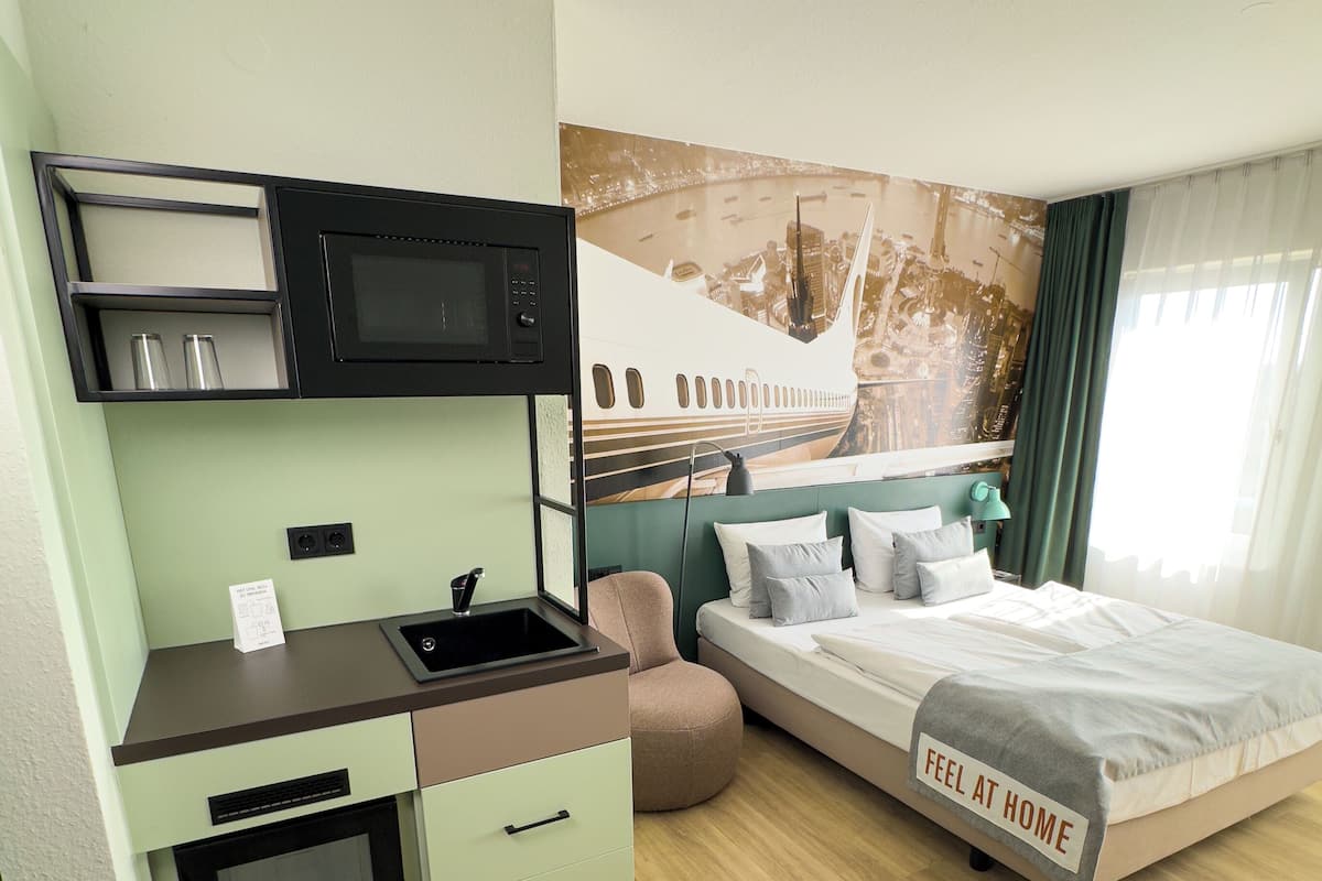 Ein kompaktes Hotelzimmer mit Küchenzeile, einem Bett mit der Aufschrift 'FEEL AT HOME', einer Stadtlandschaftsmalerei und einem Vorhangfenster.