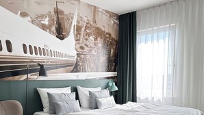 A modern bedroom with a bed, a chair, a mural of a cityscape, and a 'FEEL AT HOME' sign on the bed. - Coffee Fellows Hotel Frankfurt-Langen (Langen)
